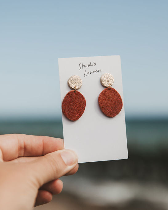 A hand holds a small white card displaying a pair of earrings against a blurred beach and sky background. The earrings have round terracotta-colored drop shapes with textured metallic gold studs above them.