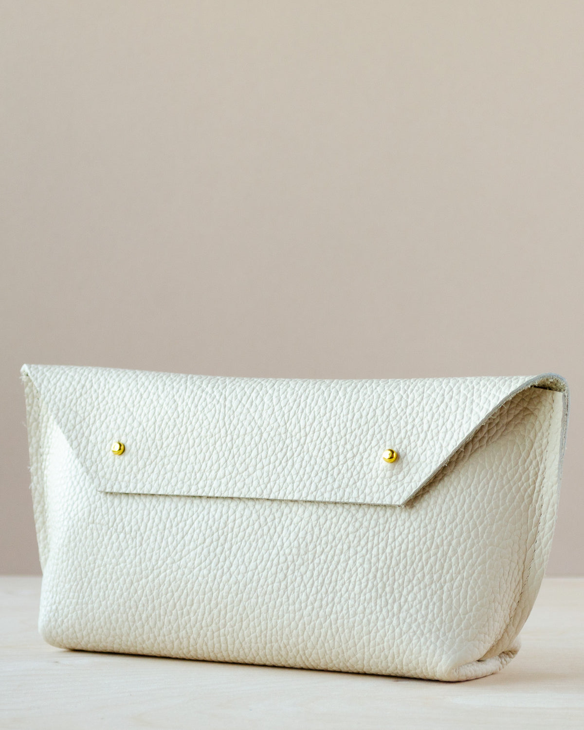 A cream-colored pebbled leather clutch bag with a flap closure and two small gold-tone metal buttons, resting on a light wooden surface against a solid beige background.