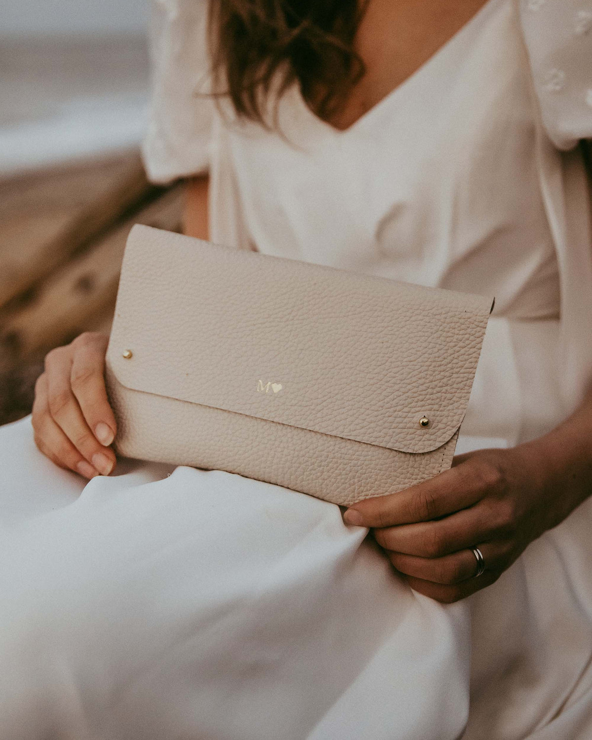 A bride in a white wedding dress holds a light beige or cream pebbled leather clutch bag with gold-tone hardware.