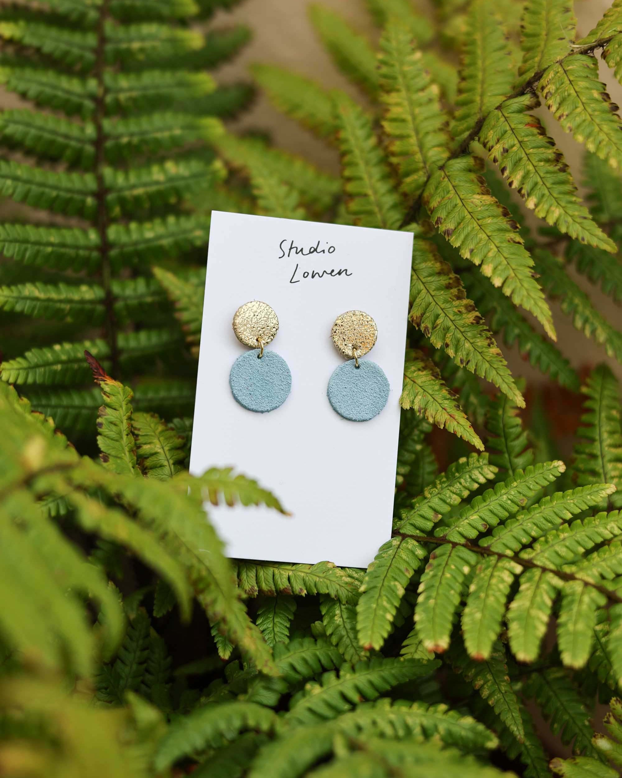 Pair of round suede and leather earrings on a card with 'Studio Lowen' branding, set against a background of green fern leaves.