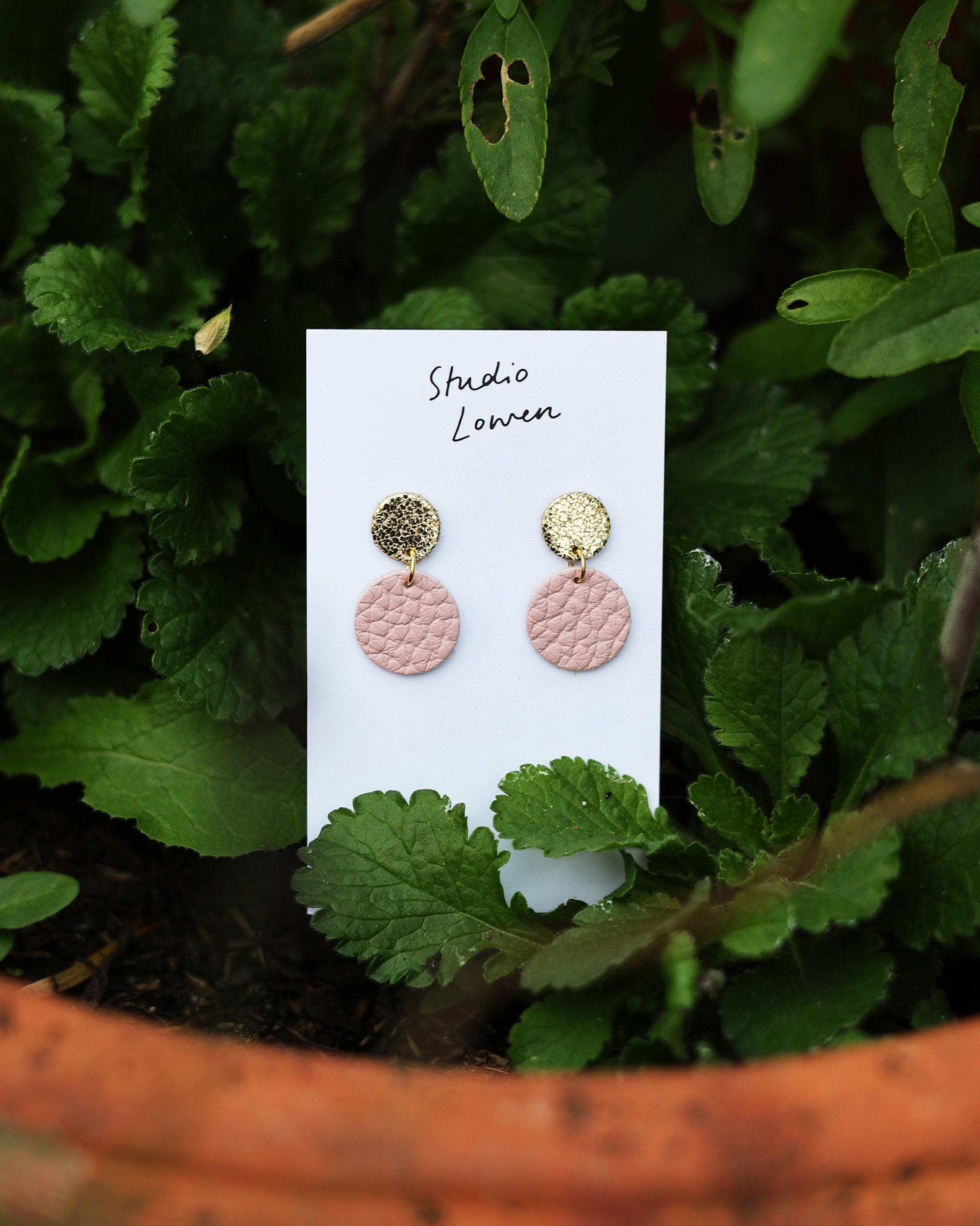 Pair of pink and gold earrings on a white card with 'Studio Lowen' branding, placed on green leaves.