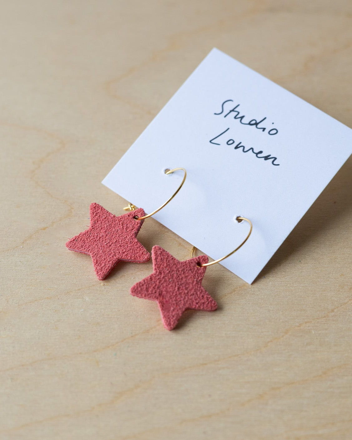 Pink star-shaped earrings on a white card with 'Studio Lowen' text, placed on a wooden surface.