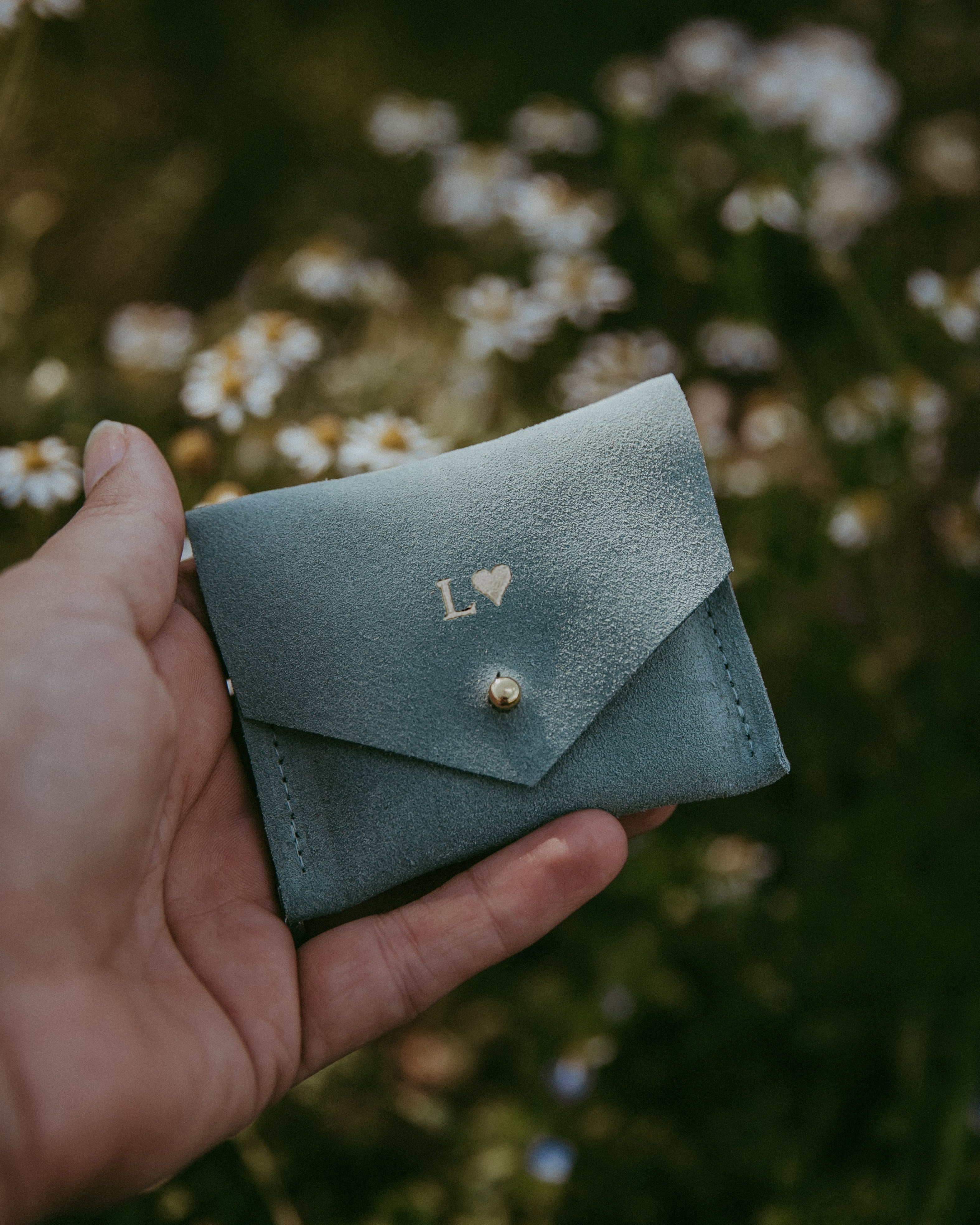 Mint green suede jewellery pouch in hands in front of daisies in summer.