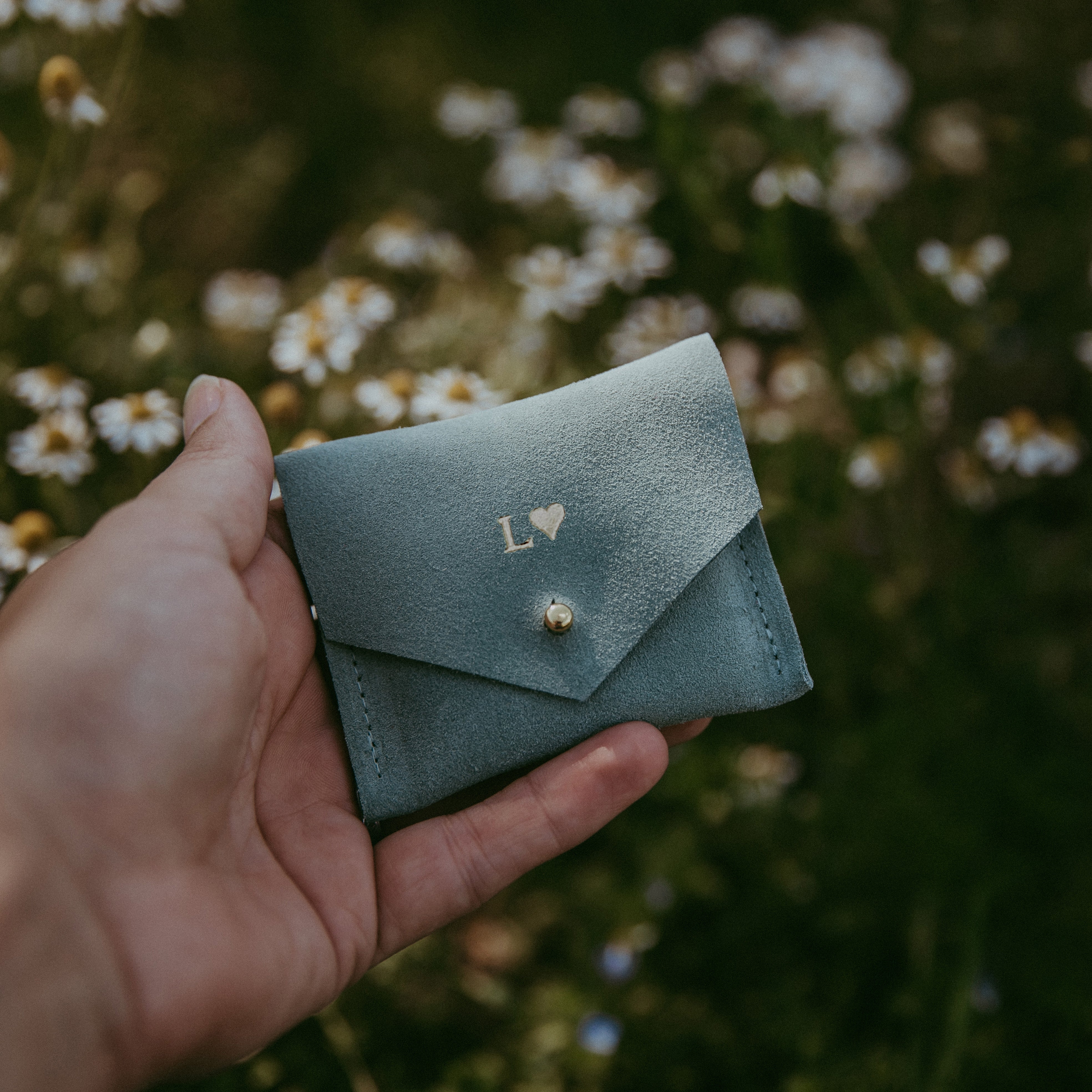 Mint green suede jewellery pouch in hands in front of daisies in summer.