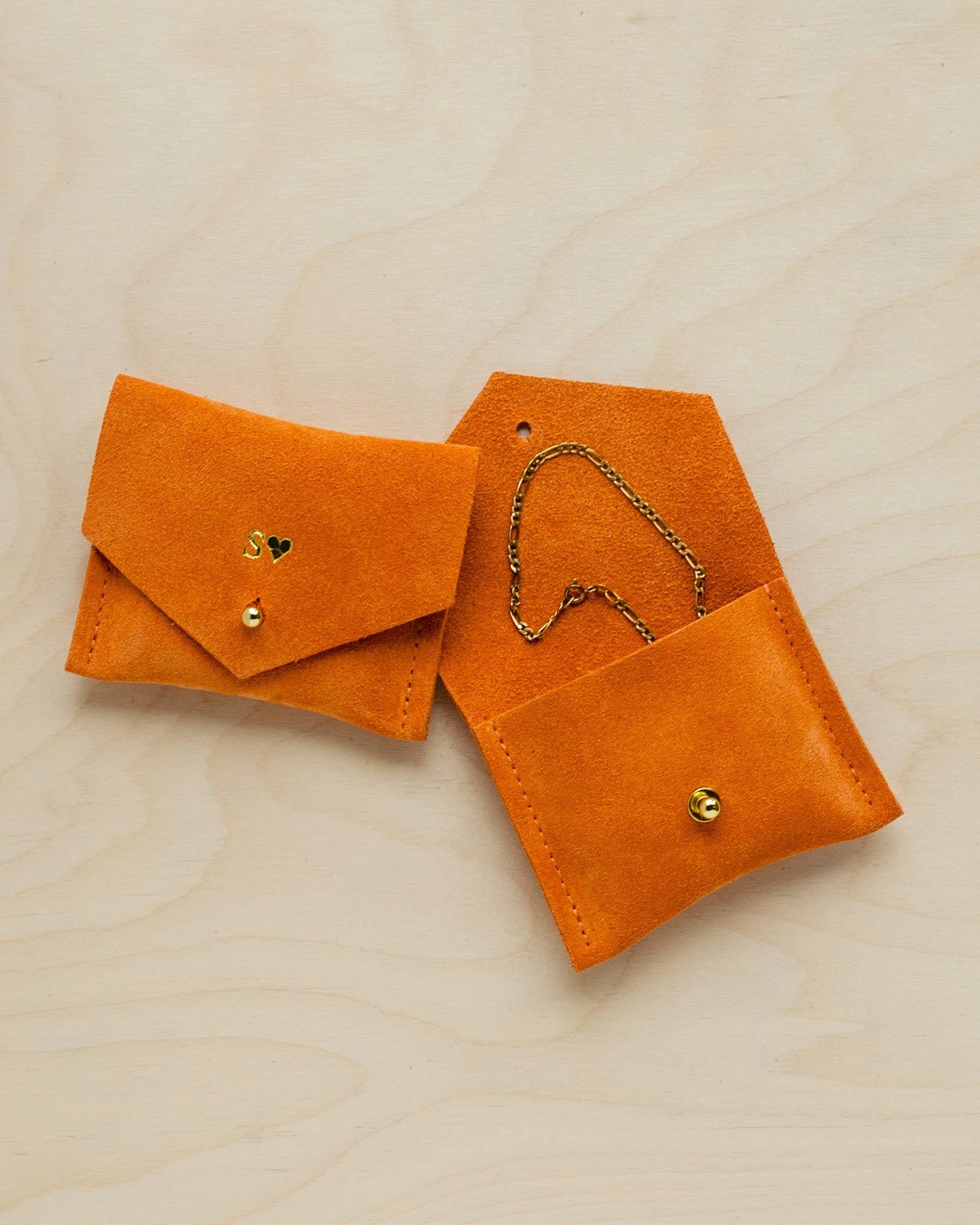 Pair of orange suede jewellery pouches sit on wooden table. One pouch bears the initial S while the other is open and displays a bracelet.