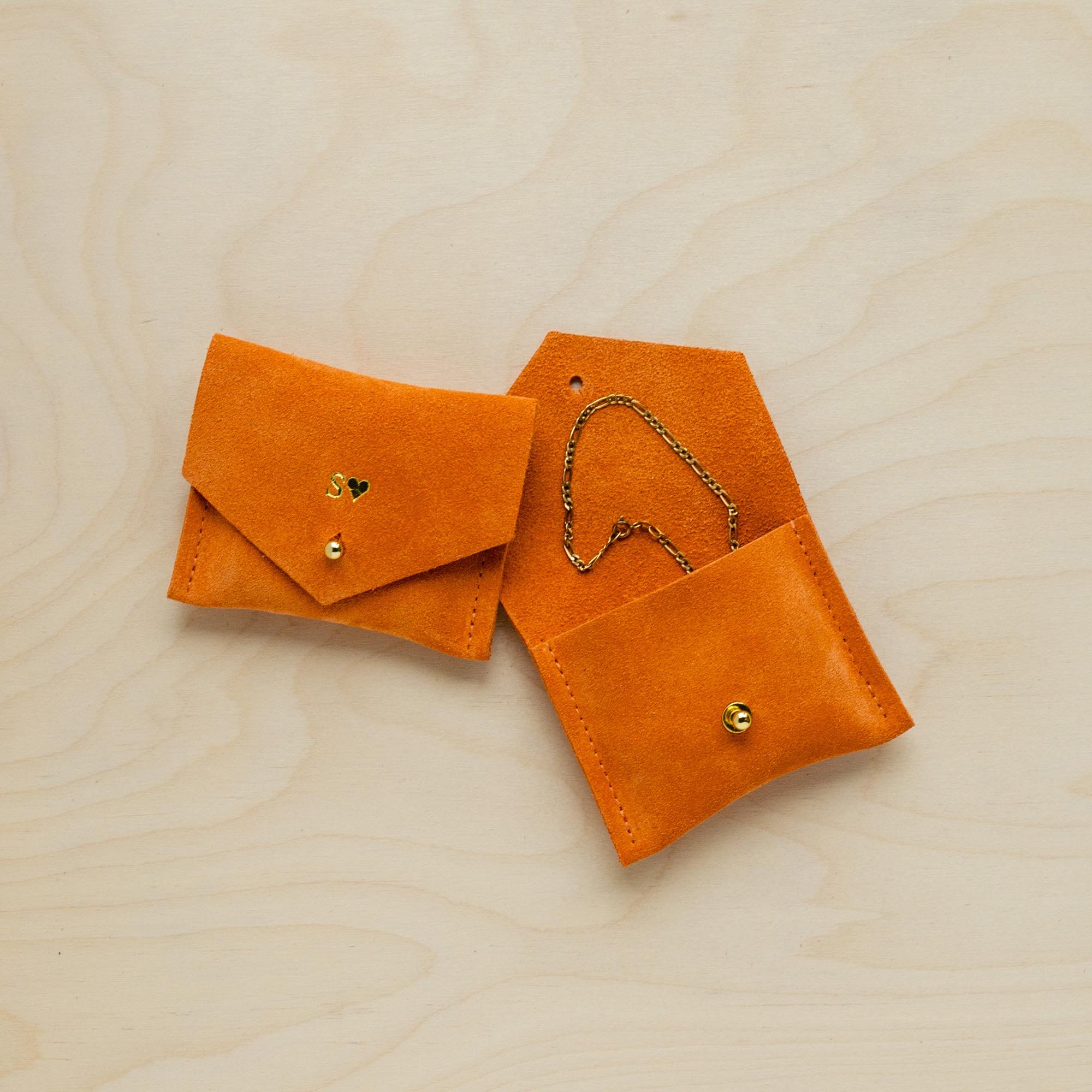 Pair of orange suede jewellery pouches sit on wooden table. One pouch bears the initial S while the other is open and displays a bracelet.