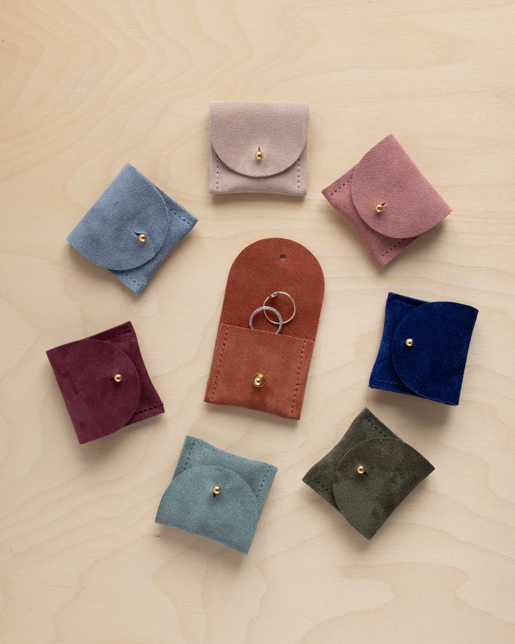 Seven small suede ring pouches in assorted colours arranged in a circle on a light wood background. The center pouch is open, holding two rings, and the others are closed with gold-tone snap buttons.
