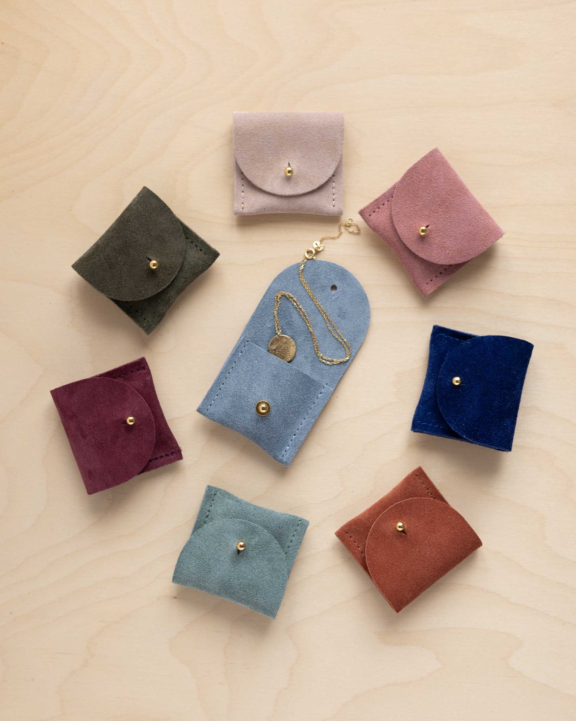 Seven small suede leather jewellery pouches in different colours arranged on a light wood background, with one blue pouch holding a gold necklace.