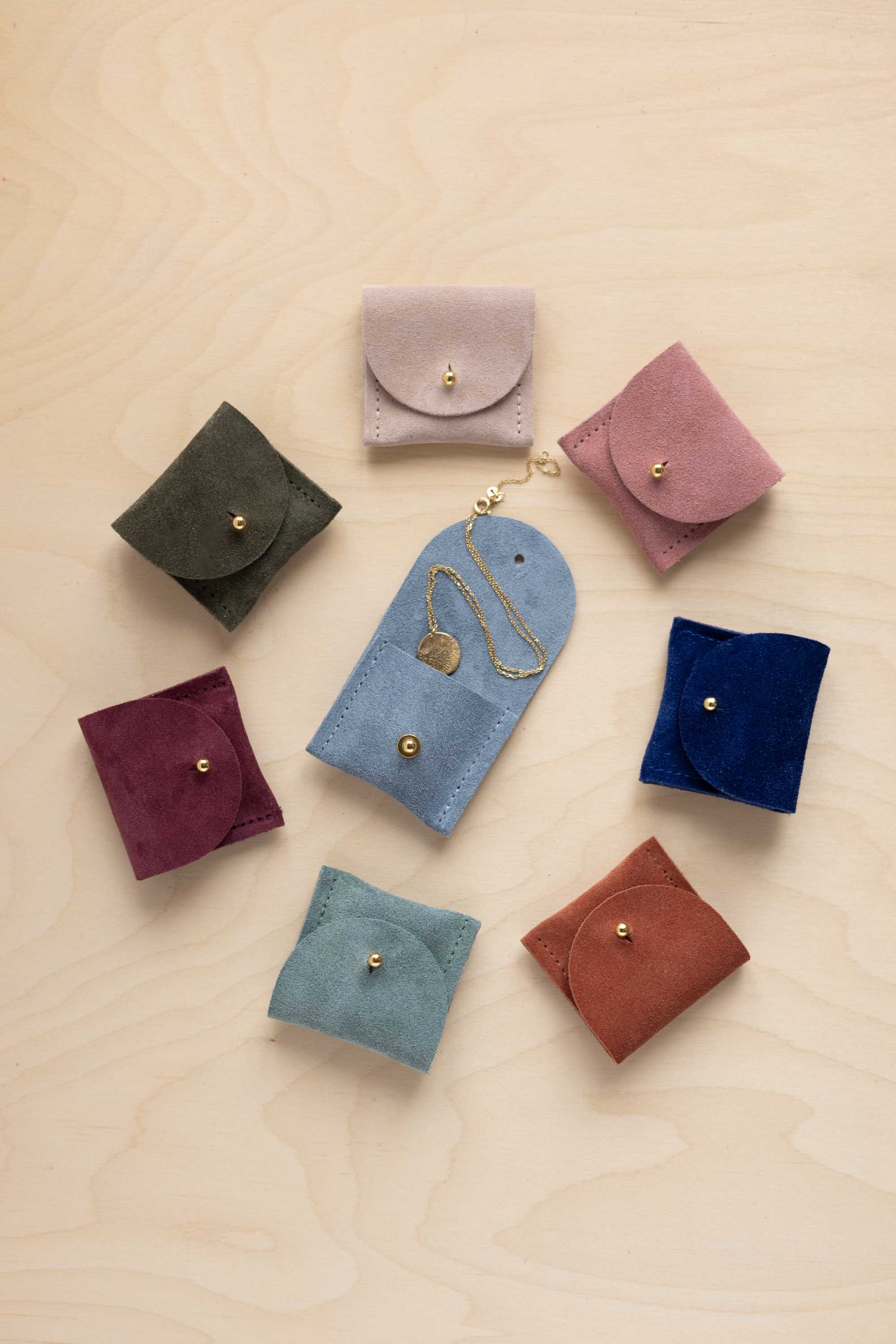 Seven small suede leather jewellery pouches in different colours arranged on a light wood background, with one blue pouch holding a gold necklace.