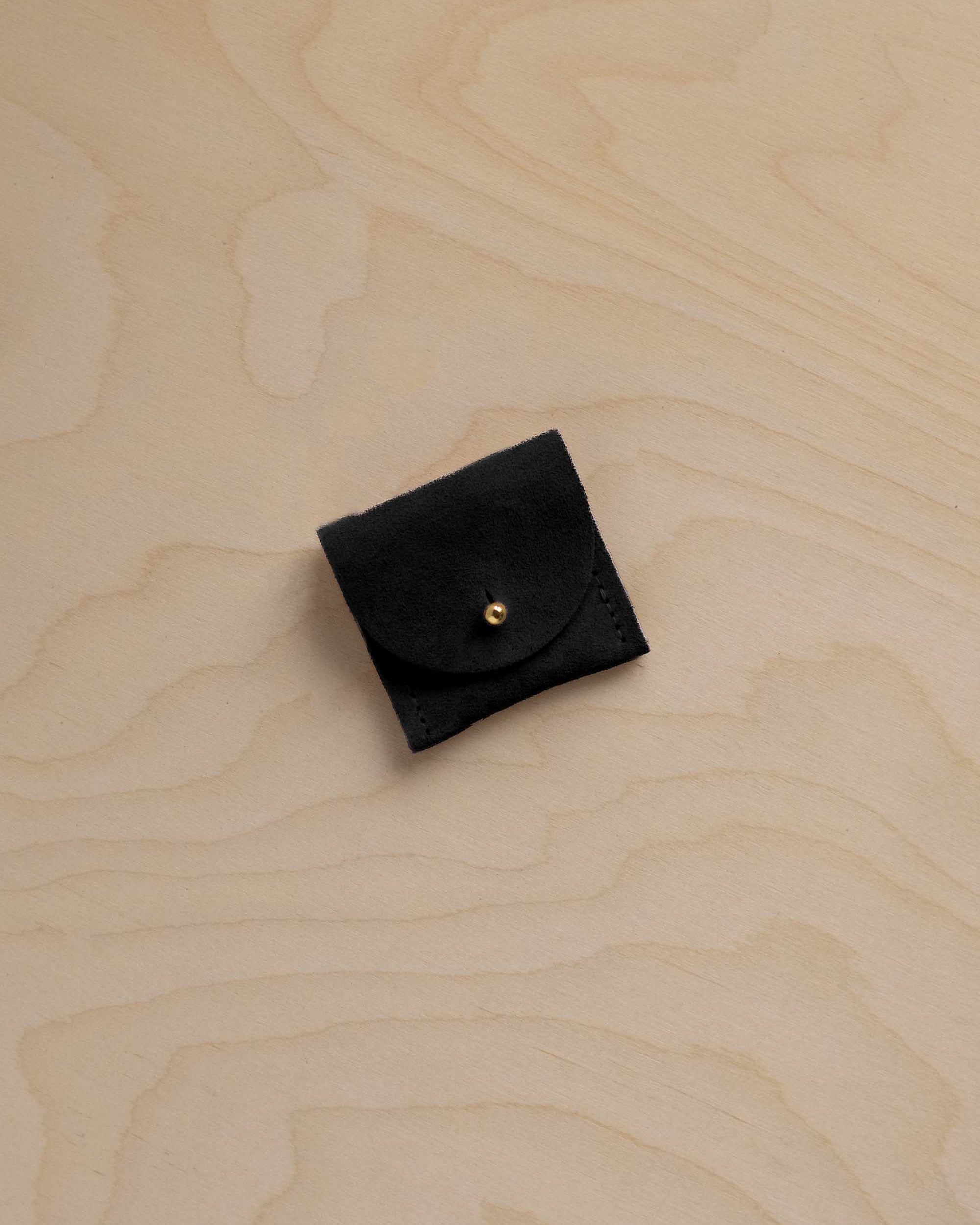 A small black suede ring pouch with a gold snap button, placed on a light wood grain surface.