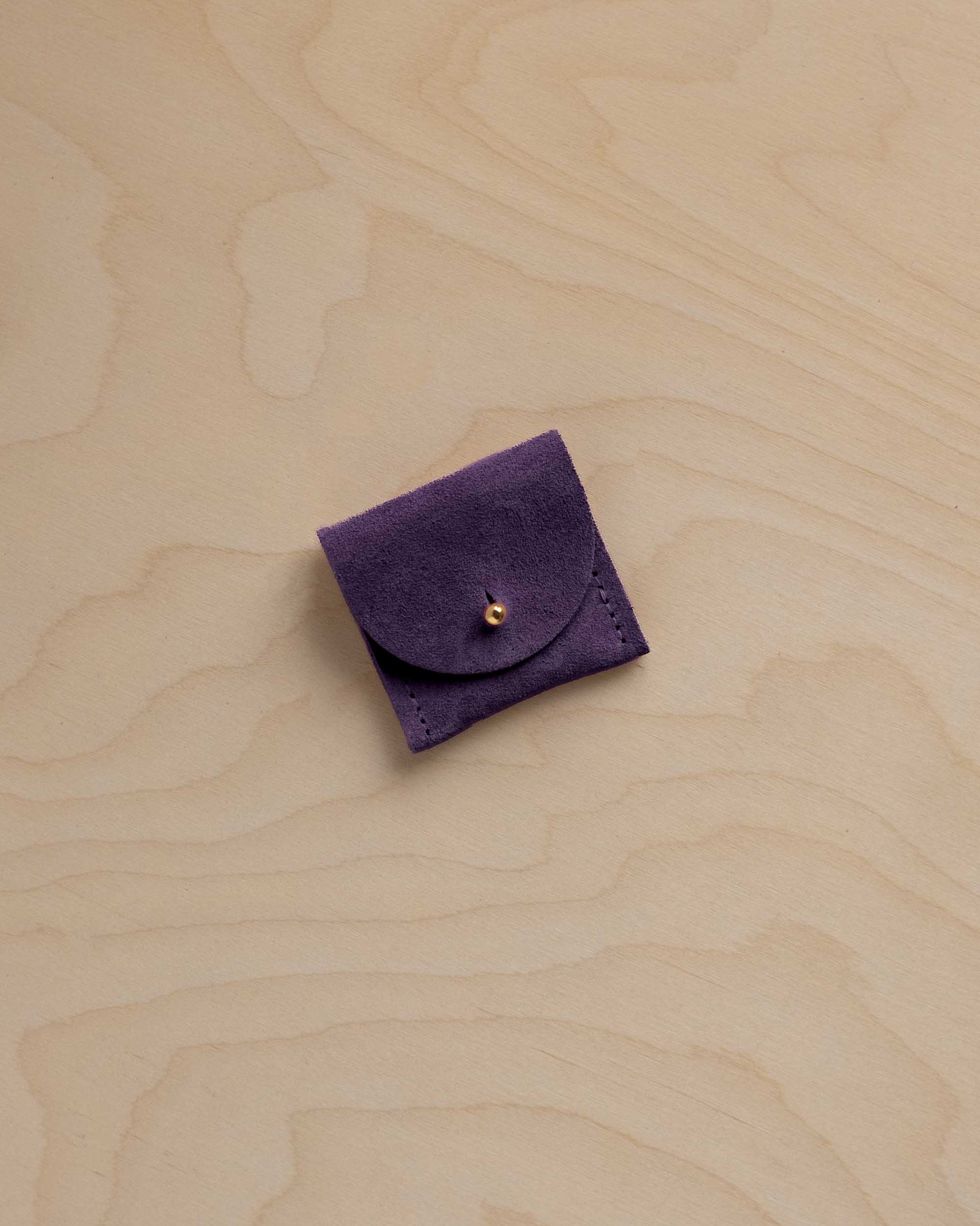 A small dark purple suede jewellery pouch with a flap closure and a gold button, placed on a light wood grain surface.