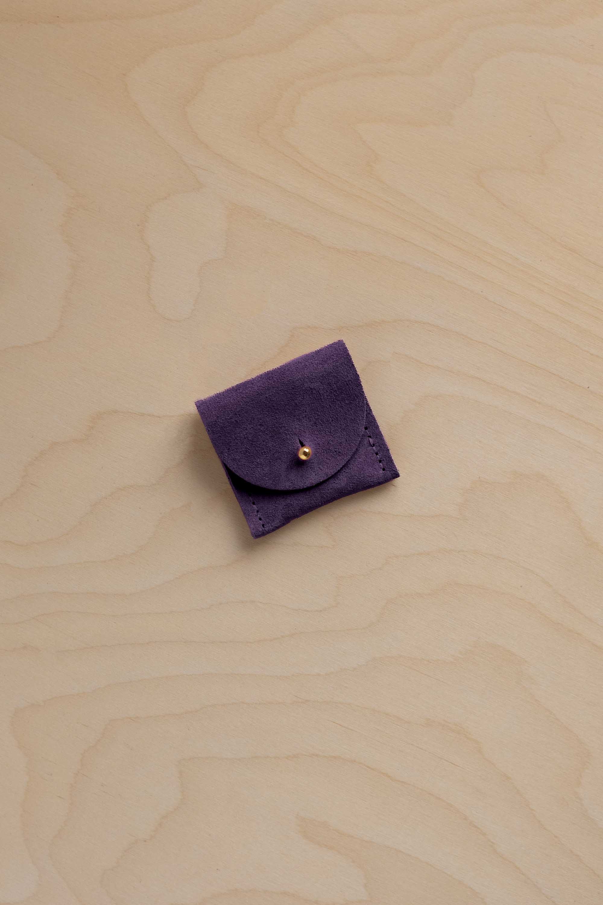 A small dark purple suede jewellery pouch with a flap closure and a gold button, placed on a light wood grain surface.