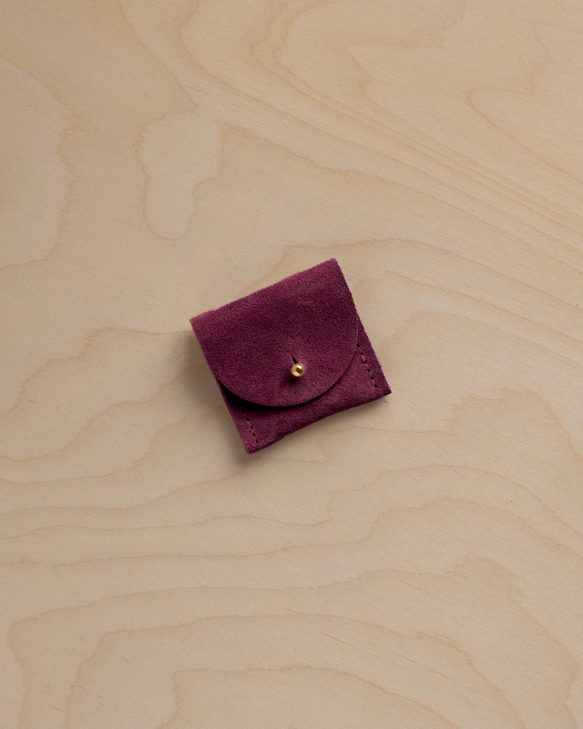 A small burgundy suede pouch with a gold button, placed on a light wood grain background.