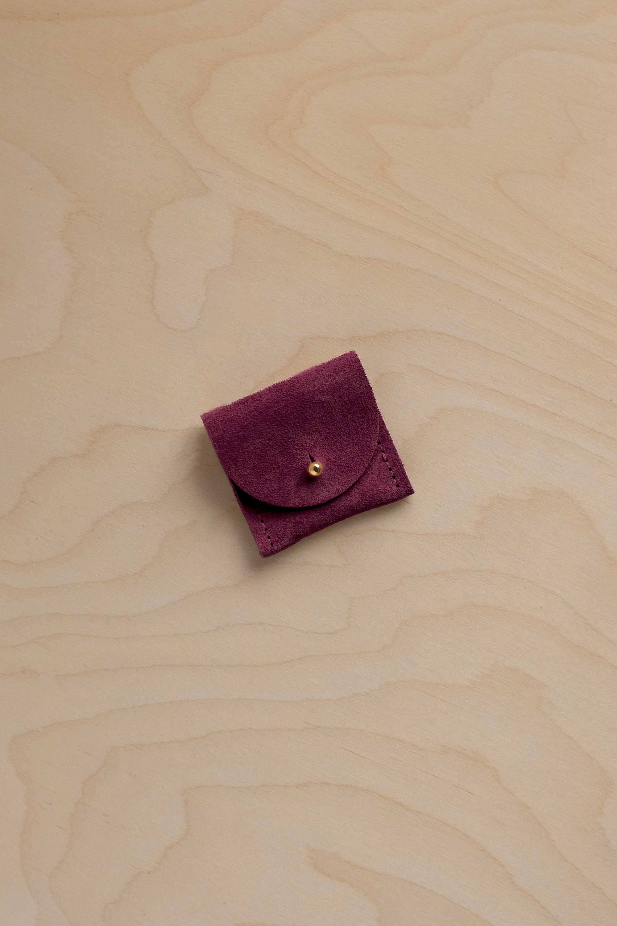 A small burgundy suede pouch with a gold button, placed on a light wood grain background.