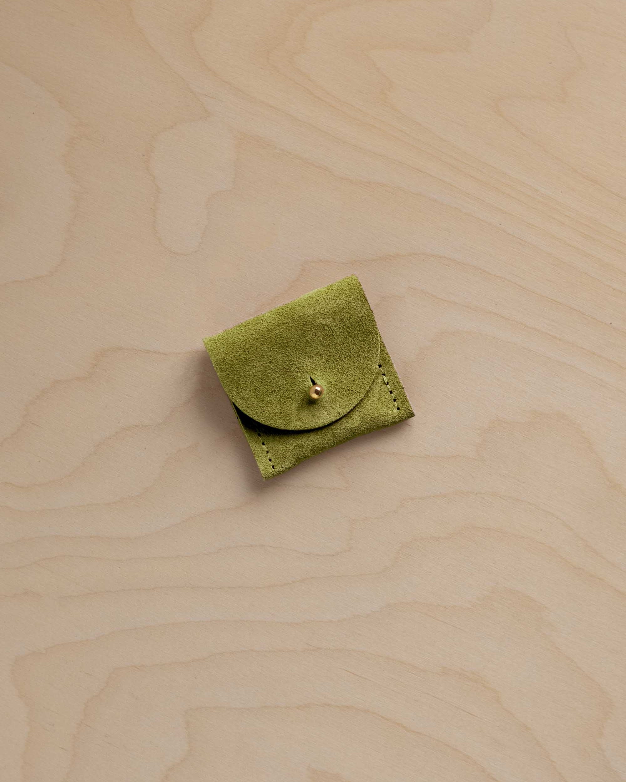 A small light olive green suede leather pouch with a button closure, placed on a light beige wood-grain surface.