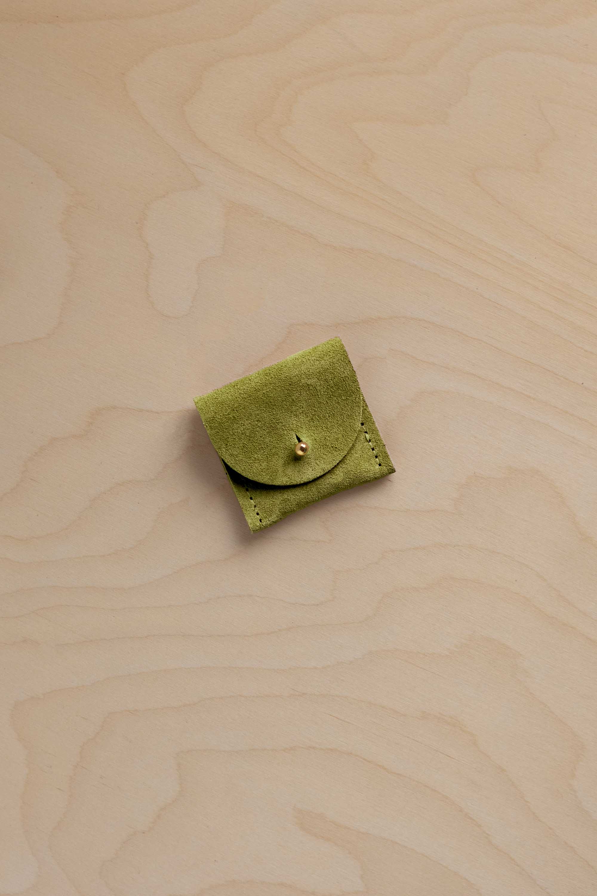 A small light olive green suede leather pouch with a button closure, placed on a light beige wood-grain surface.
