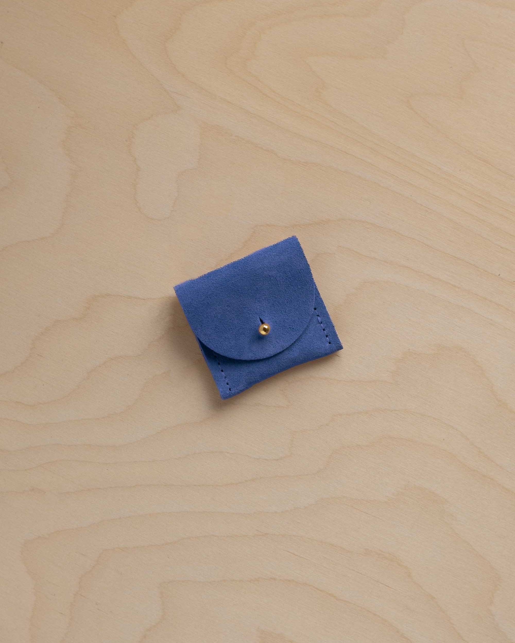 A small cobalt blue leather pouch with a flap closure and a tiny gold button, placed on a light wood grain surface.
