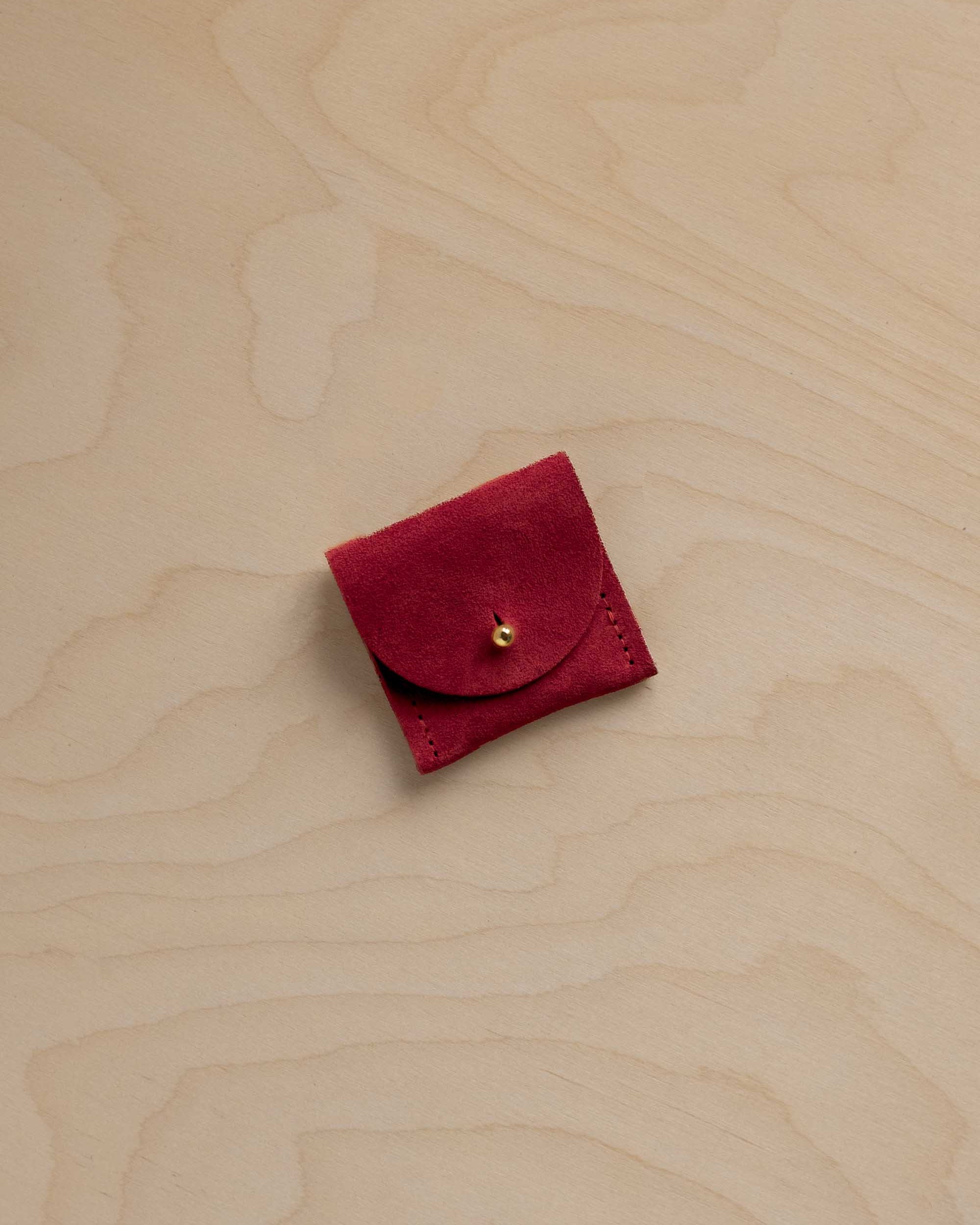 A small deep red suede leather pouch with a gold button closure, placed on a light beige wood grain background.
