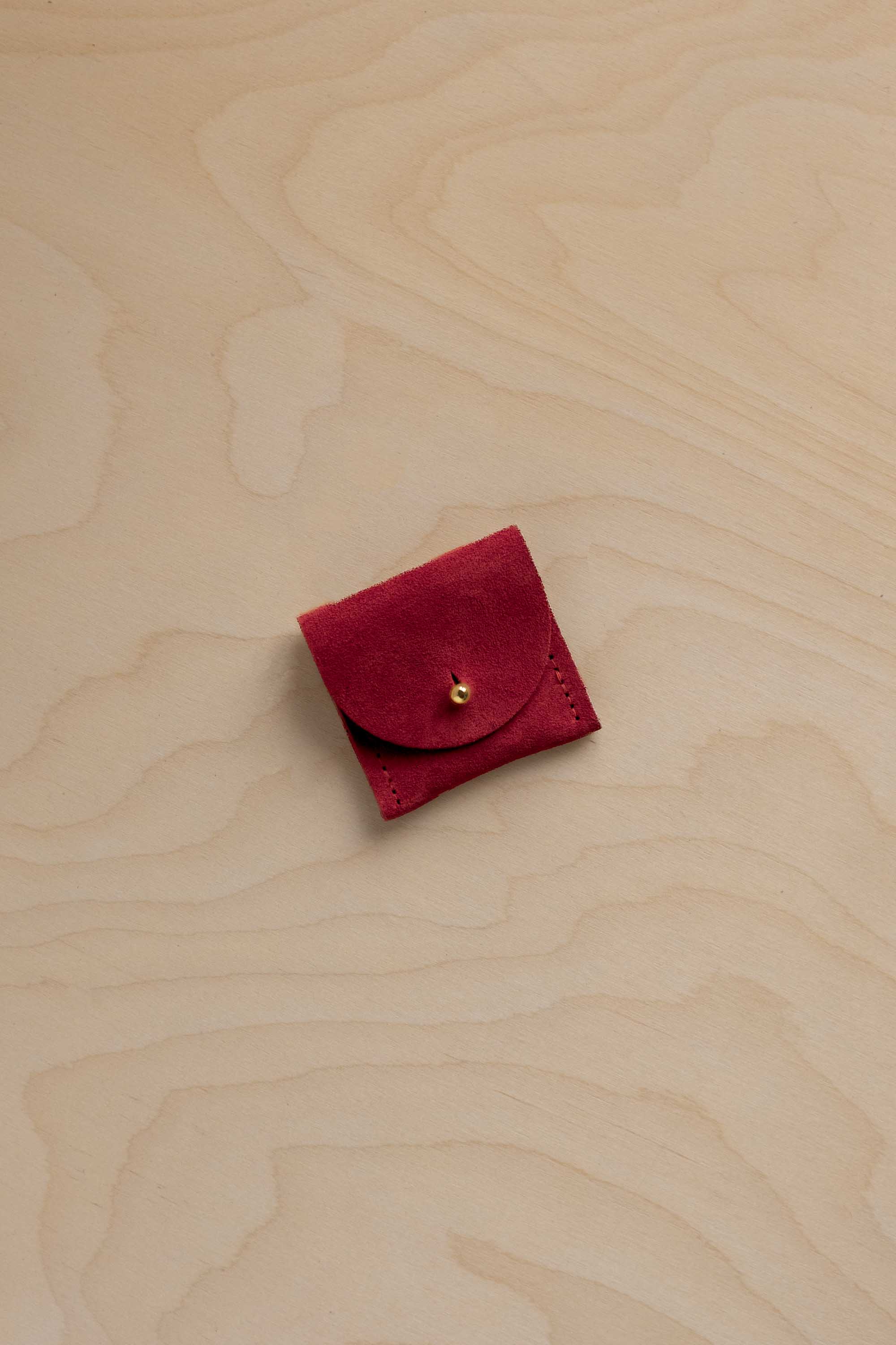 A small deep red suede leather pouch with a gold button closure, placed on a light beige wood grain background.