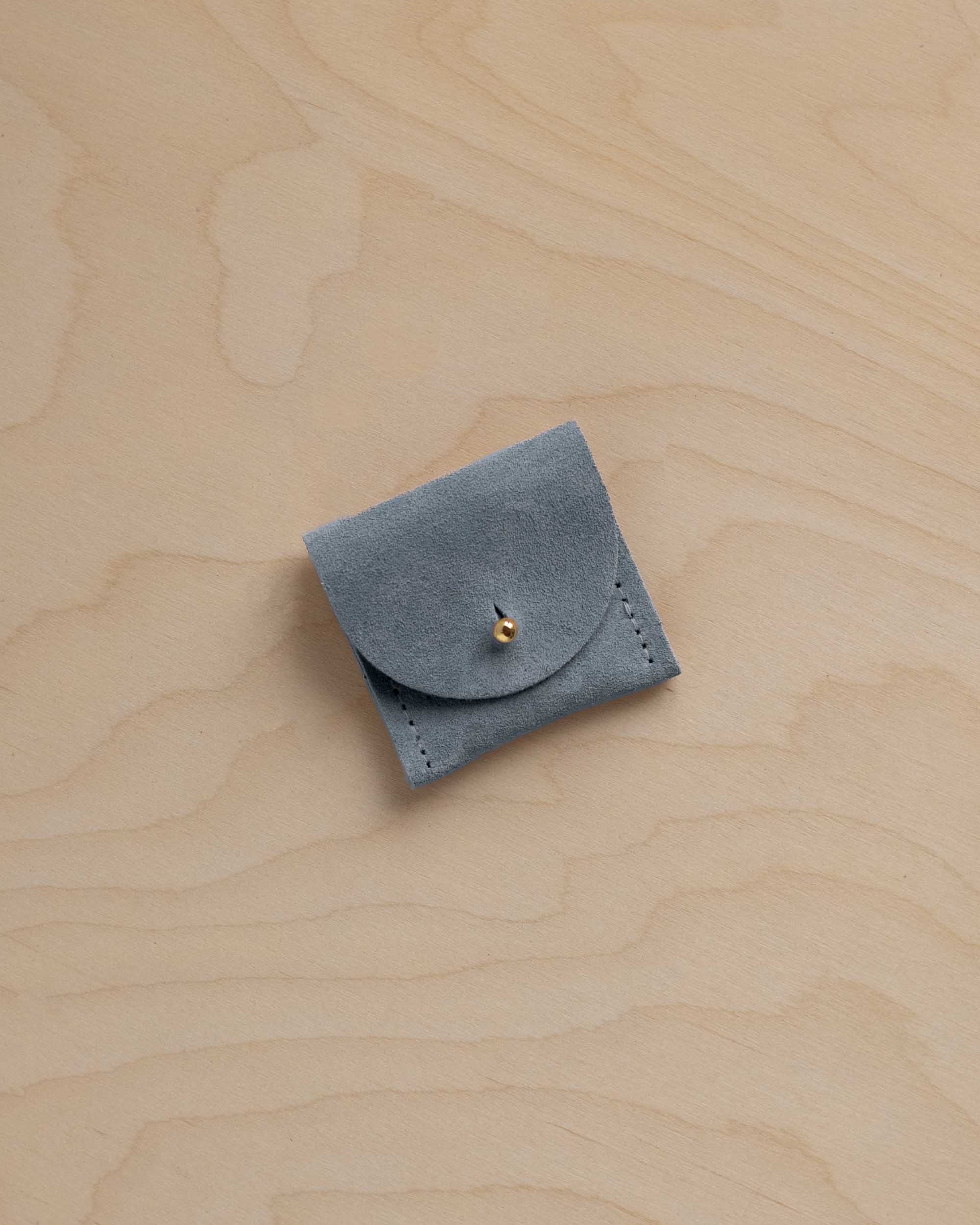 A small blue gray suede ring pouch with a flap closure and a tiny gold button, placed on a light wood grain surface.