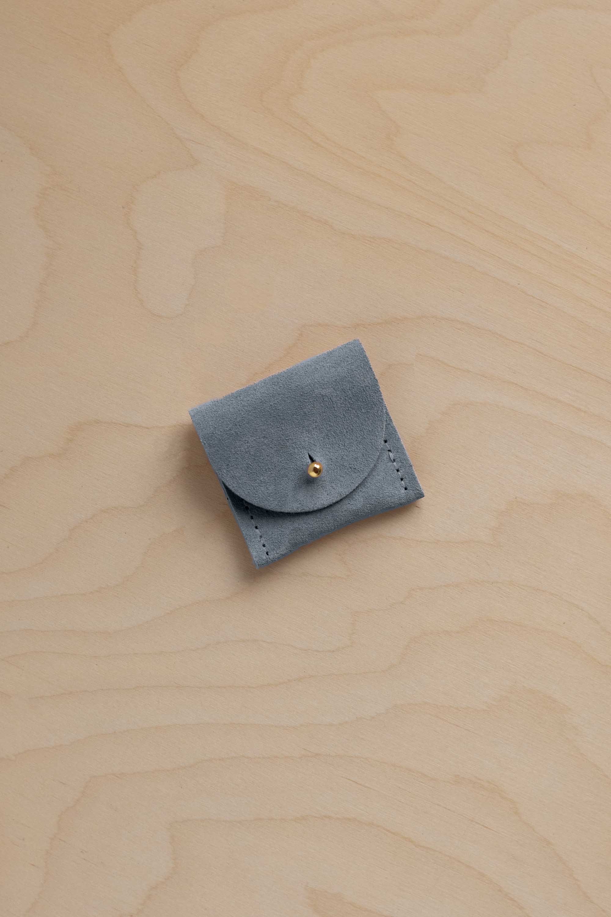 A small blue gray suede ring pouch with a flap closure and a tiny gold button, placed on a light wood grain surface.