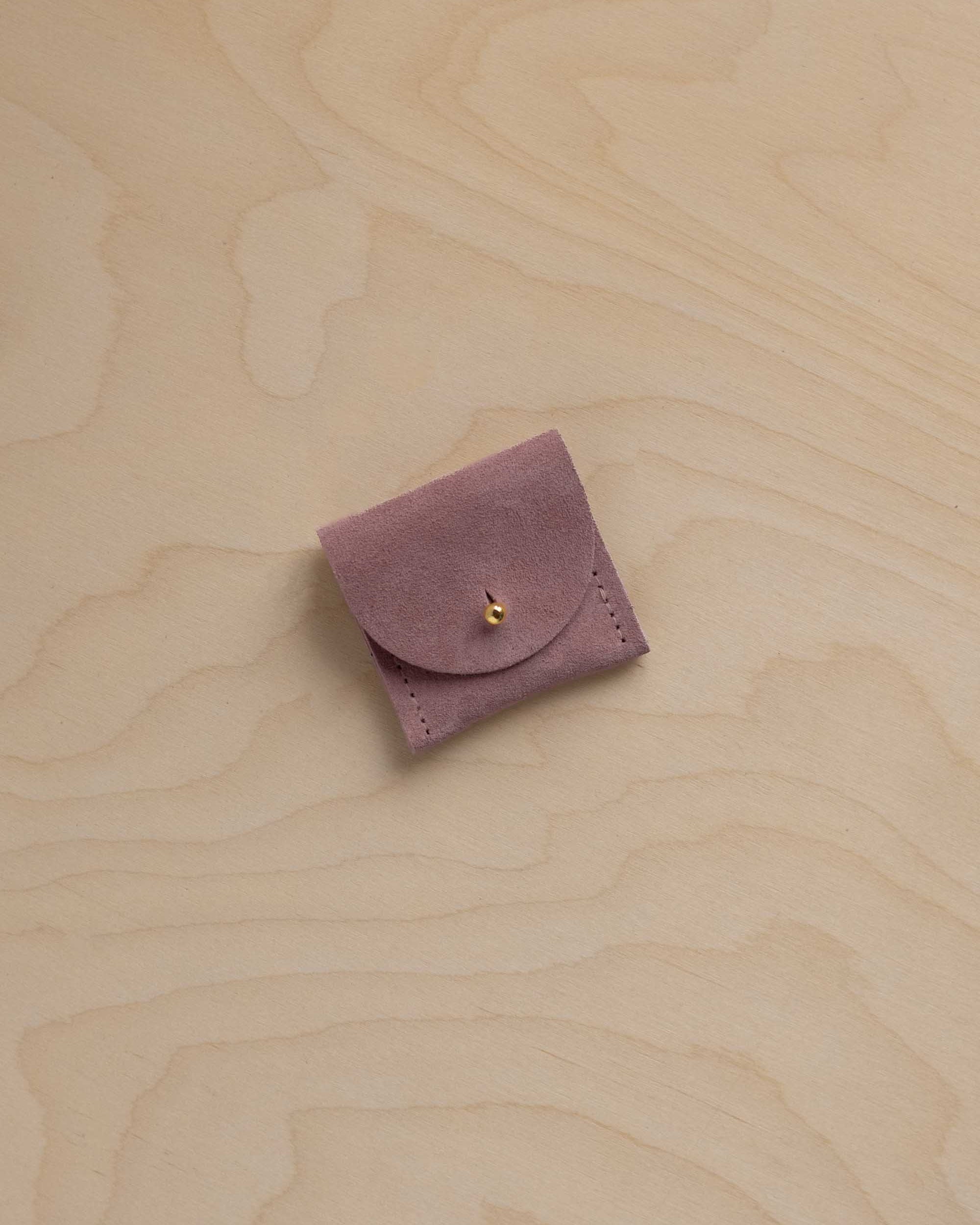 A small dusty mauve purple suede leather pouch with a flap closure and a tiny gold fastening, placed on a light wood grain surface.