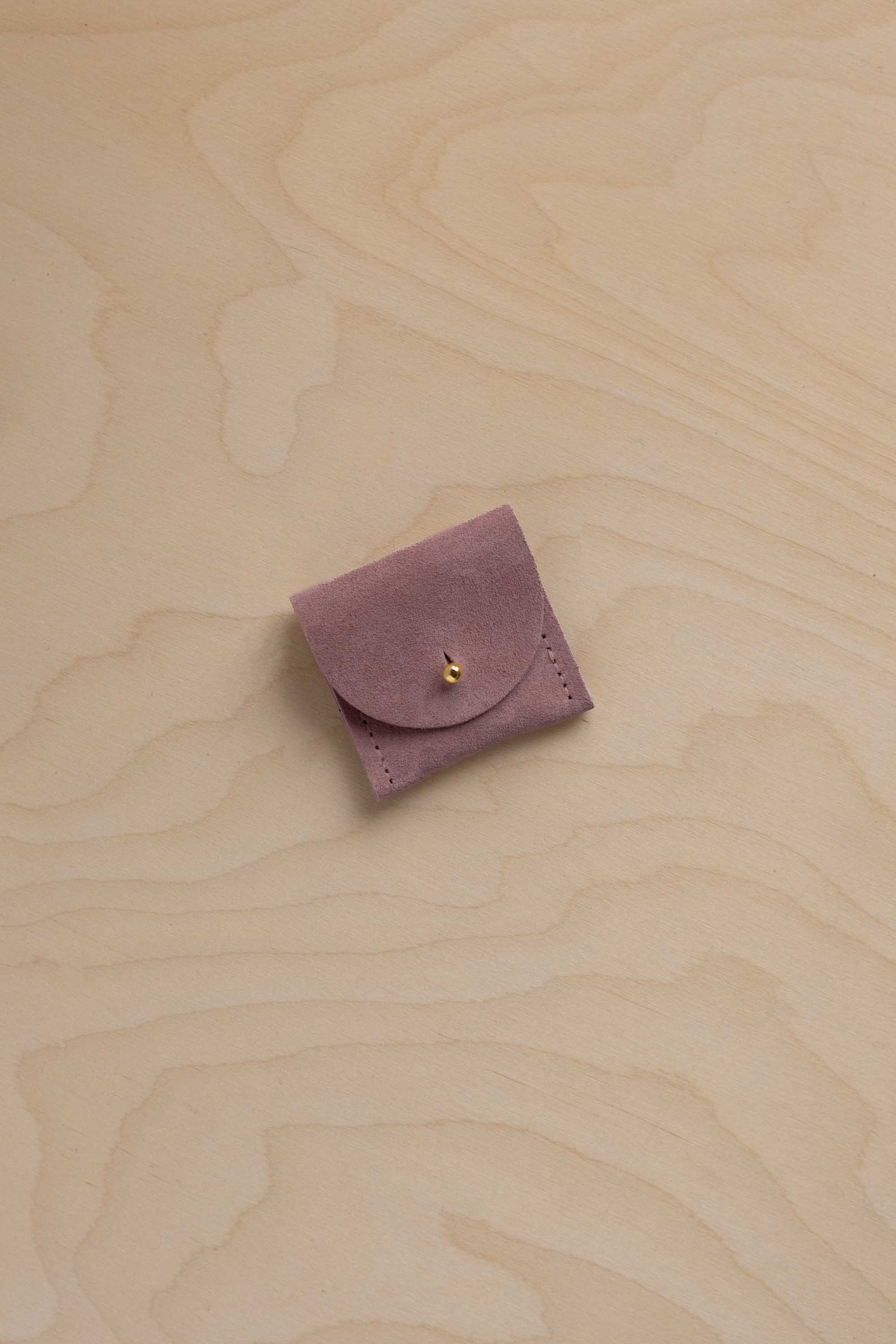 A small dusty mauve purple suede leather pouch with a flap closure and a tiny gold fastening, placed on a light wood grain surface.