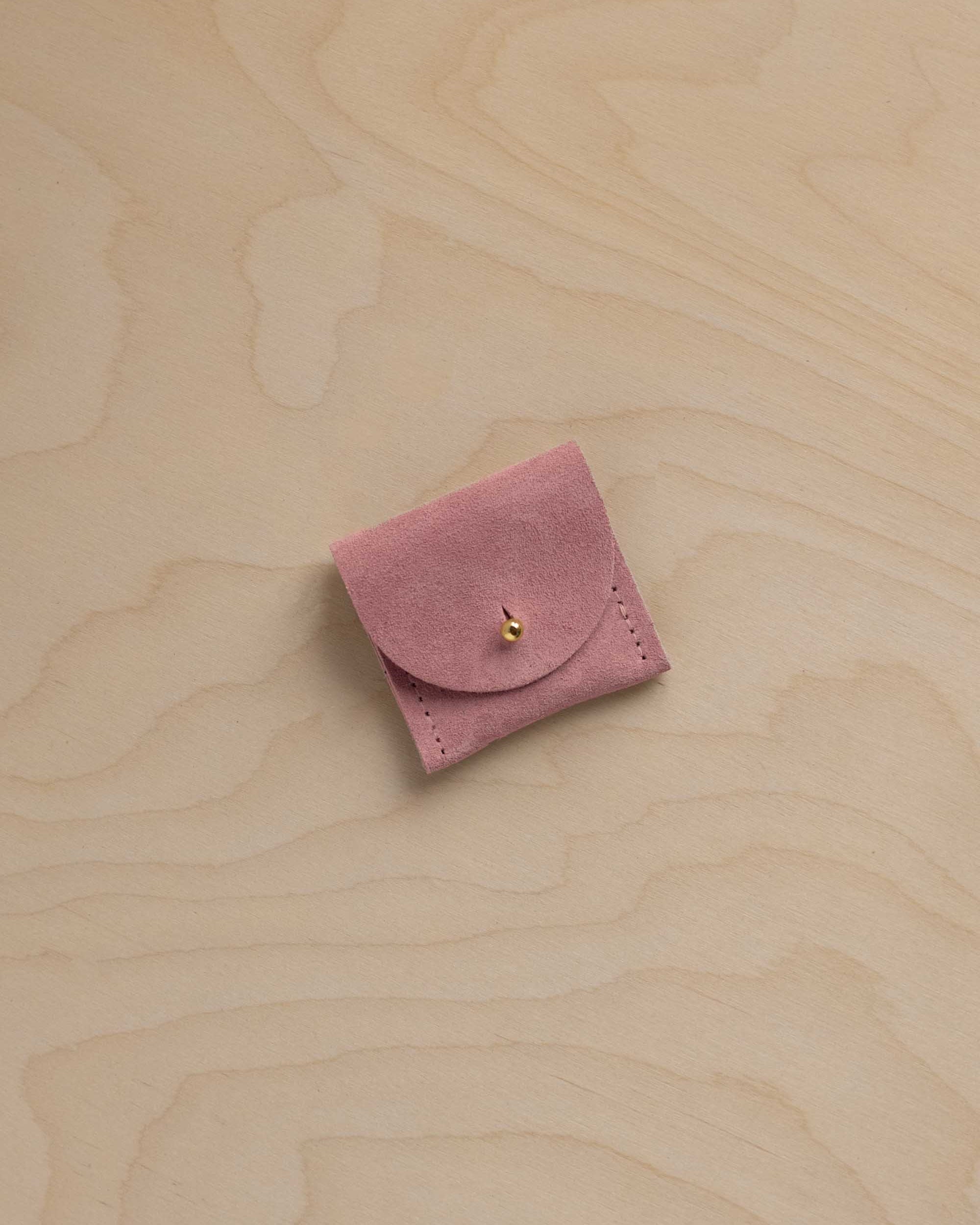 A small dusty pink suede jewelry pouch with a flap closure and a tiny gold bead accent, resting on a light wood grain surface.