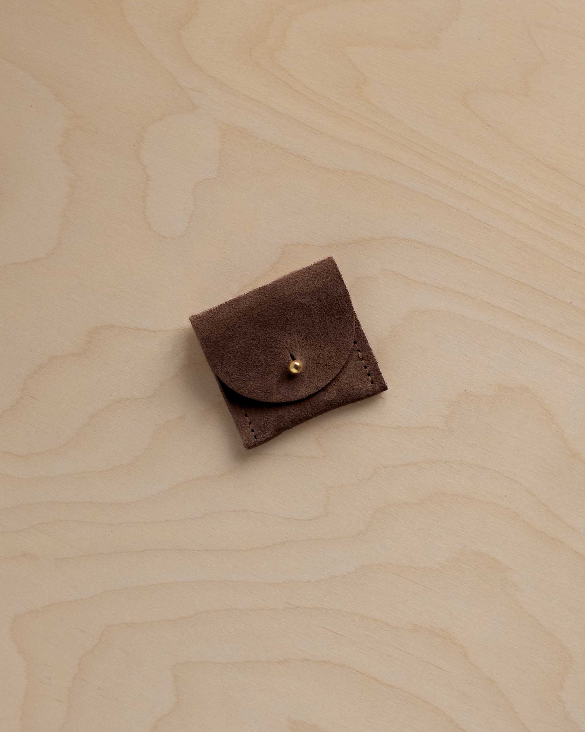 A small dark brown suede leather pouch with a flap closure and a gold snap button, placed on a light wood grain surface.