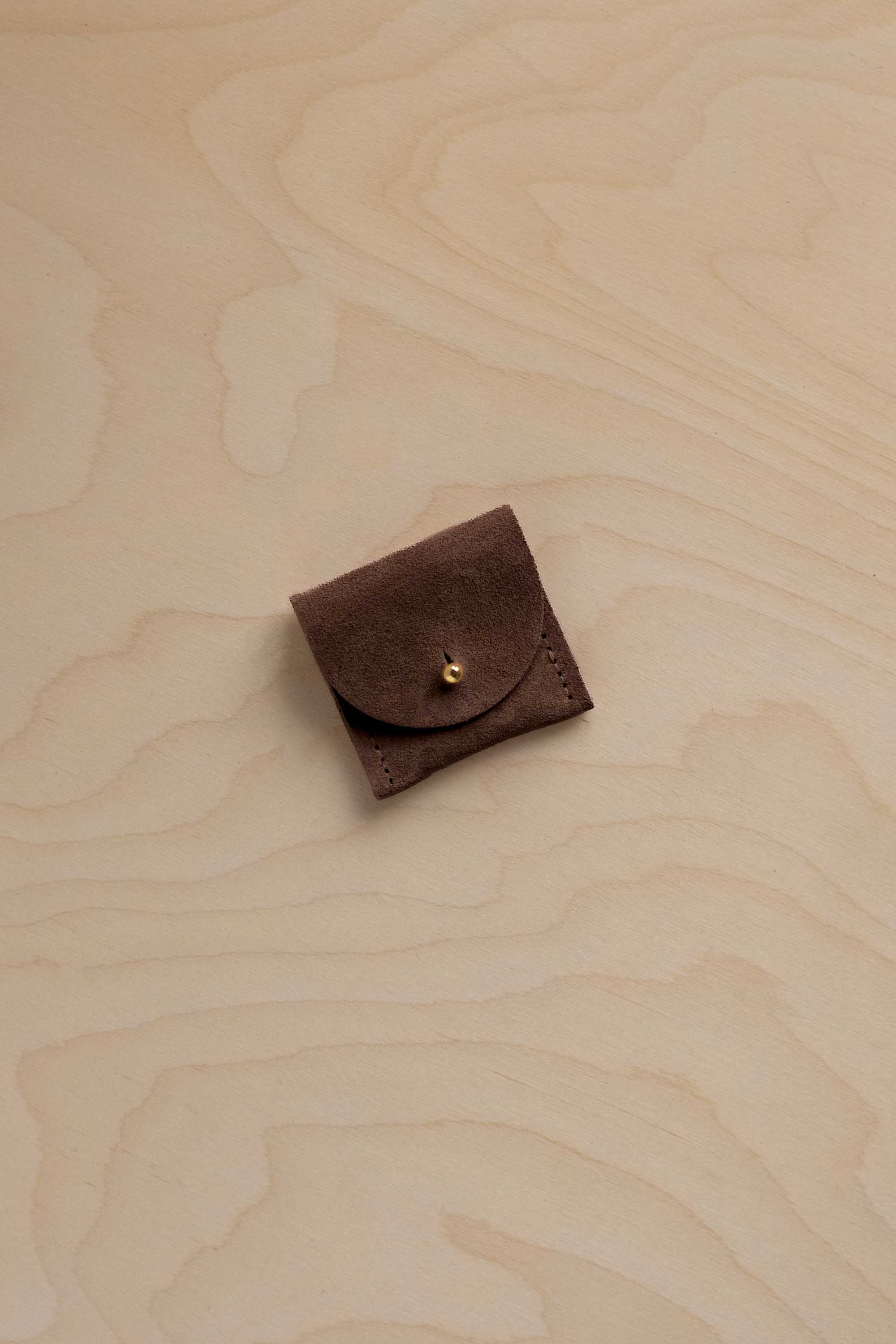 A small dark brown suede leather pouch with a flap closure and a gold snap button, placed on a light wood grain surface.