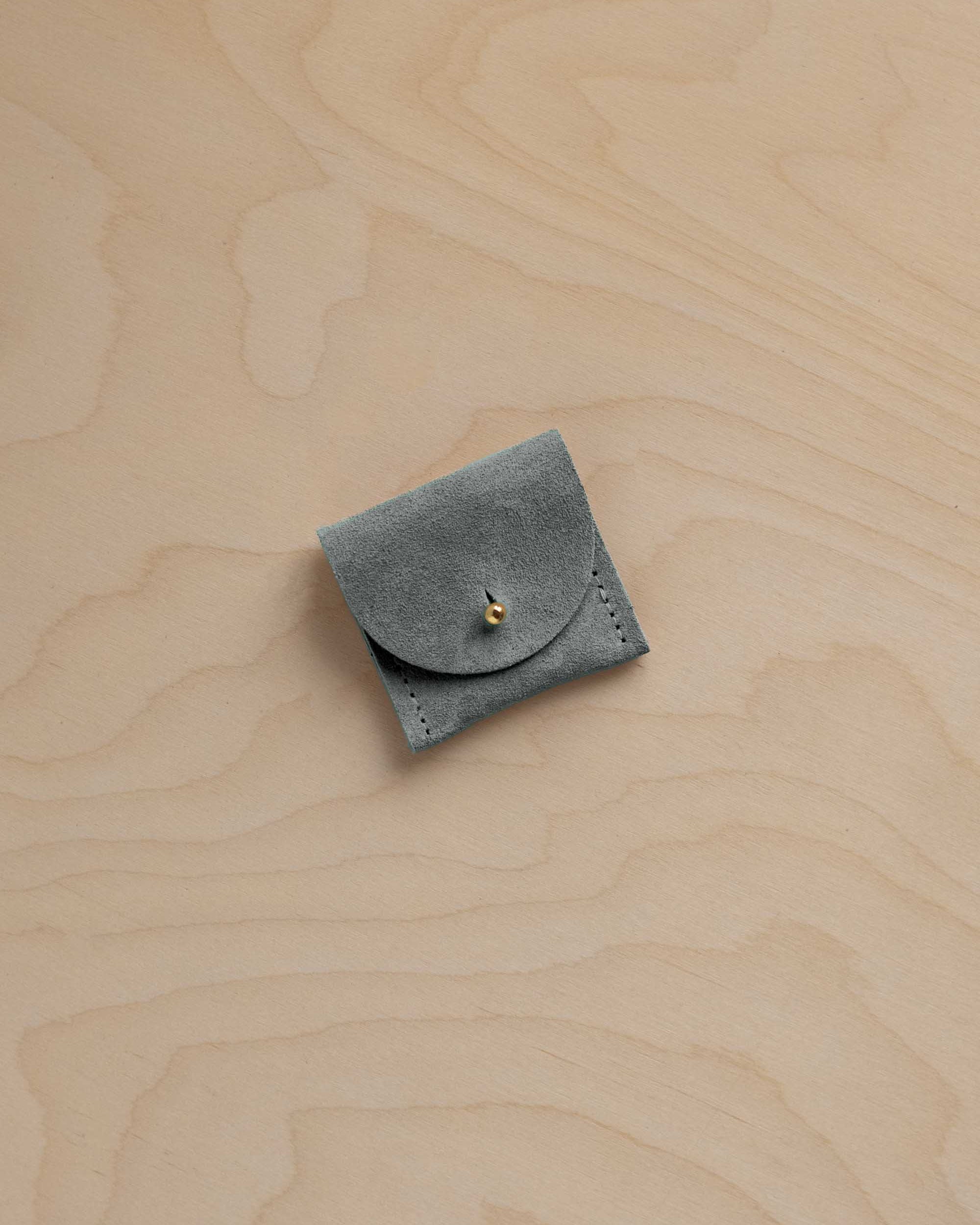 A small sage green suede pouch with a flap closure and a gold fastening, placed on a light wood grain surface.