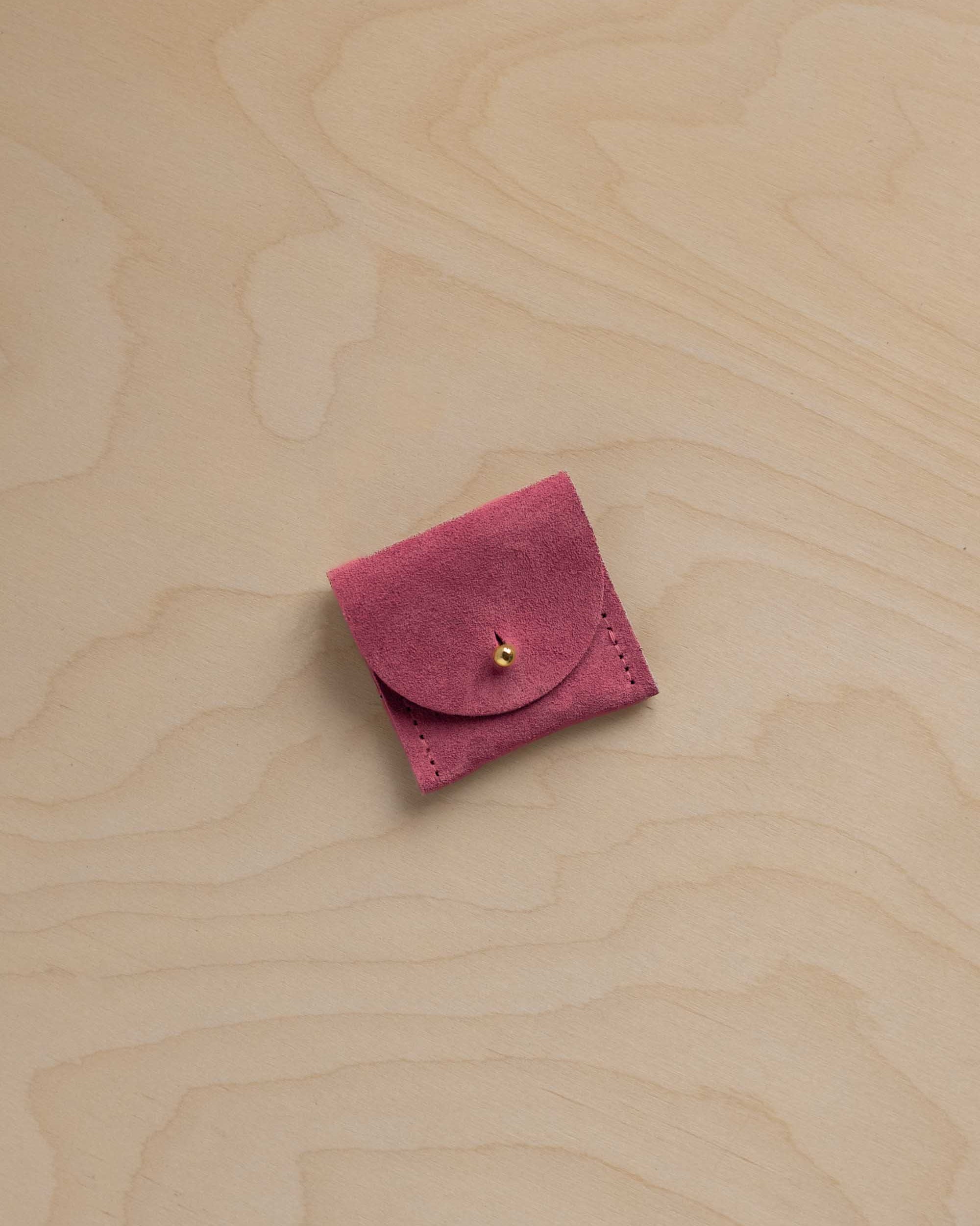 A small rose pink suede pouch with a gold button closure, placed on a light beige wood grain background.