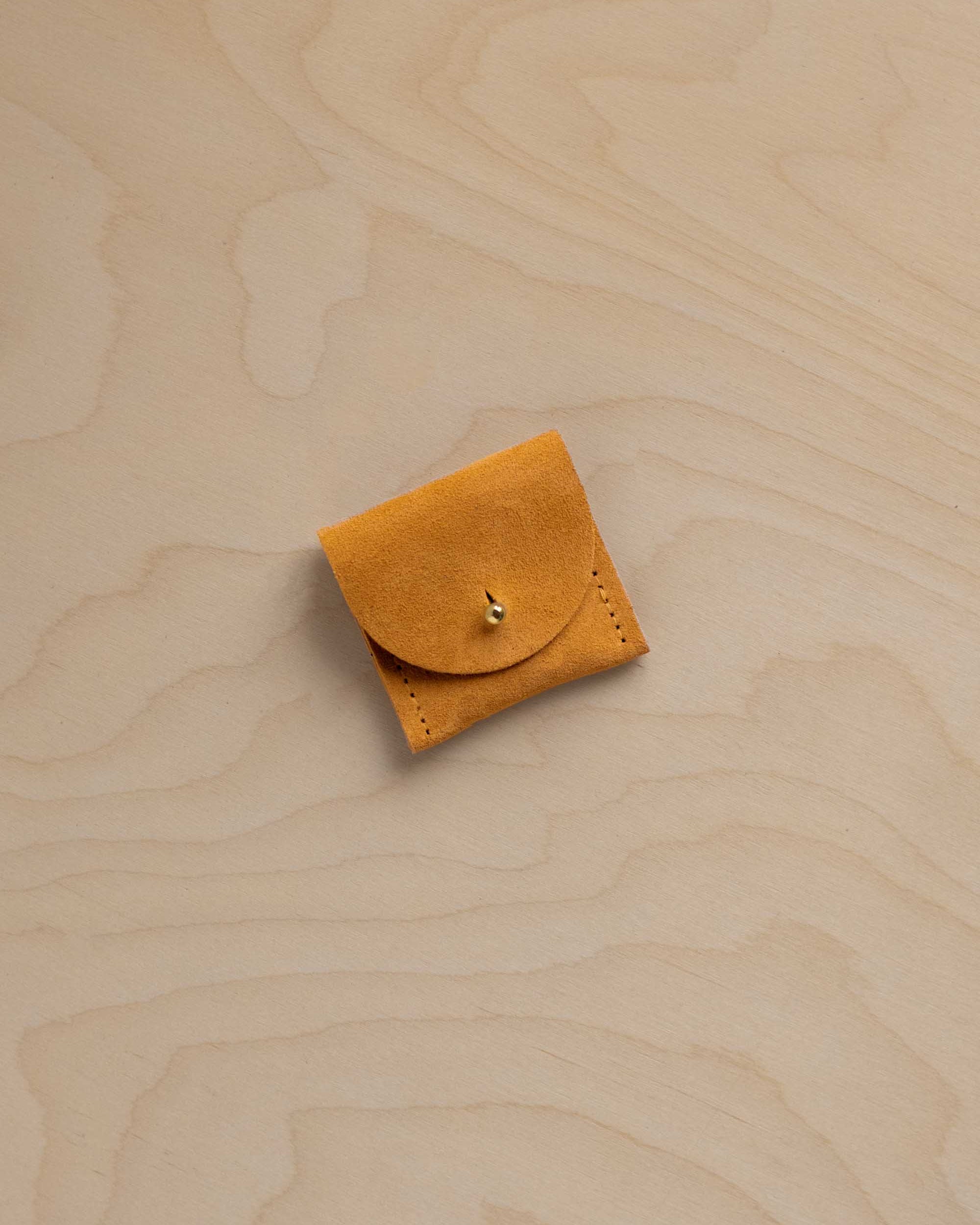 A small yellowy orange suede leather pouch with a flap closure and a gold button, placed on a light wood grain surface.