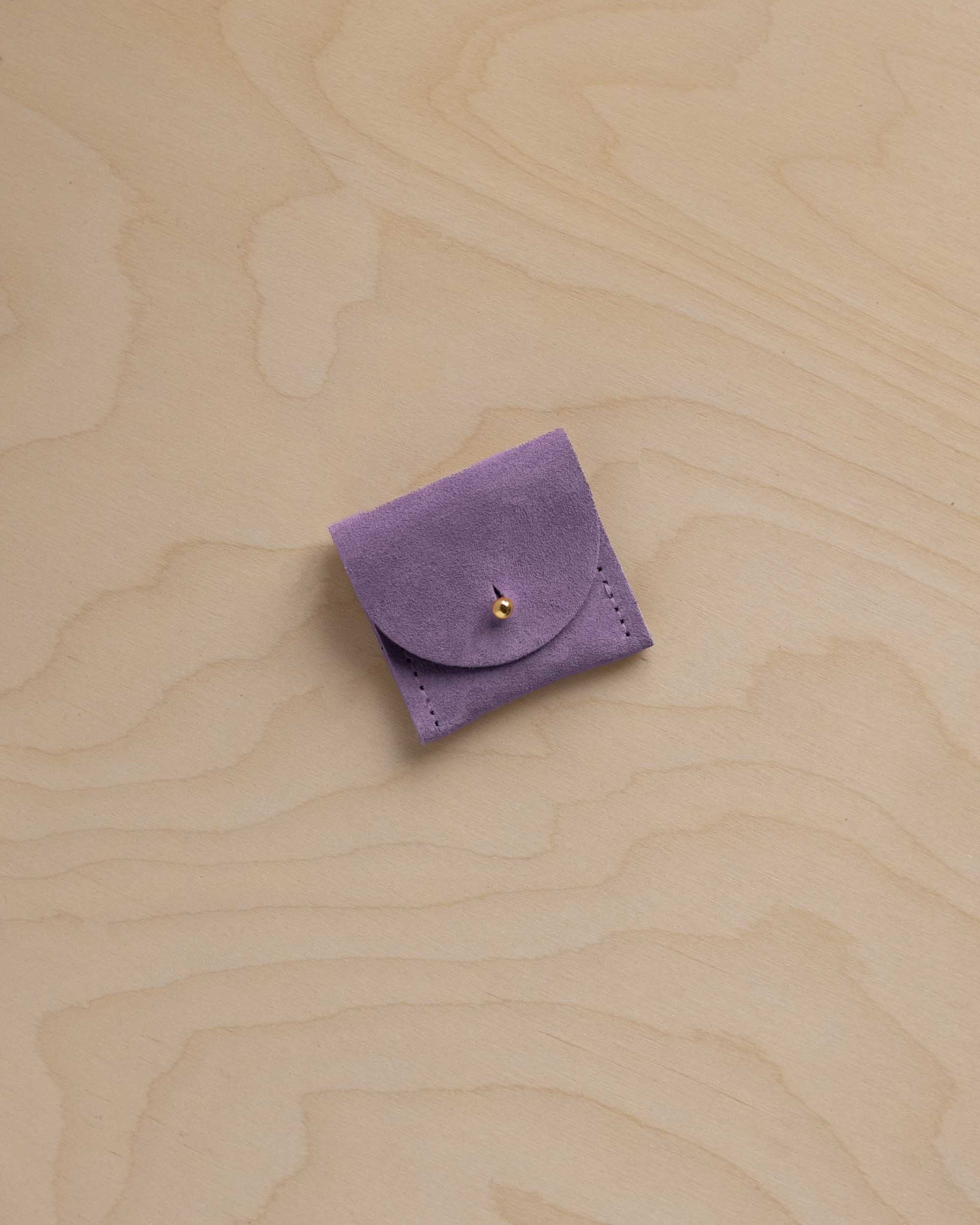A small lavender purple suede ring pouch with a flap closure and a tiny gold button, placed on a light beige wood grain surface.