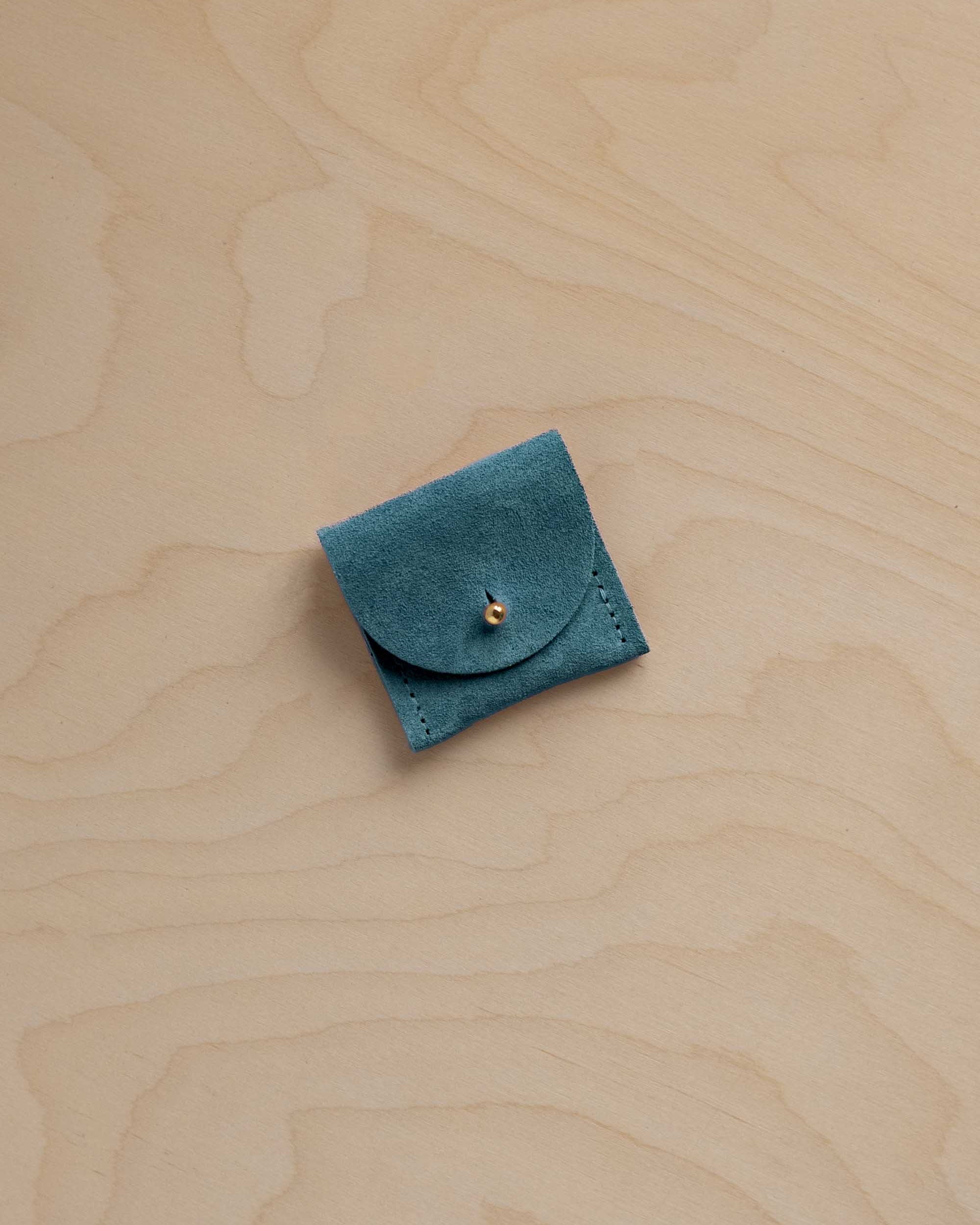 A small teal blue suede pouch with a flap closure and a tiny gold fastening, placed on a light wood grain surface.