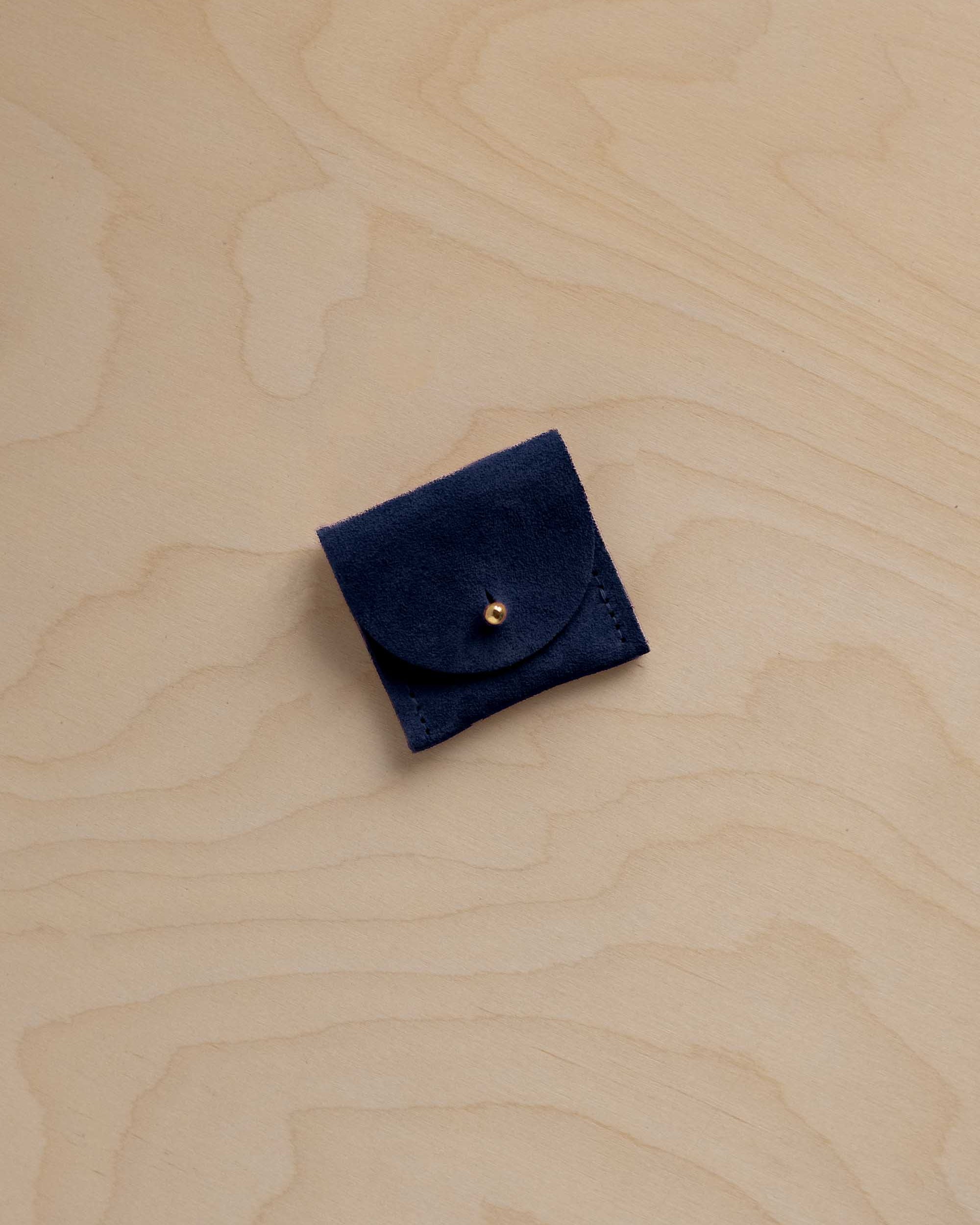 A small navy blue suede leather pouch with a flap closure and a gold-tone button, placed on a light wood grain surface.