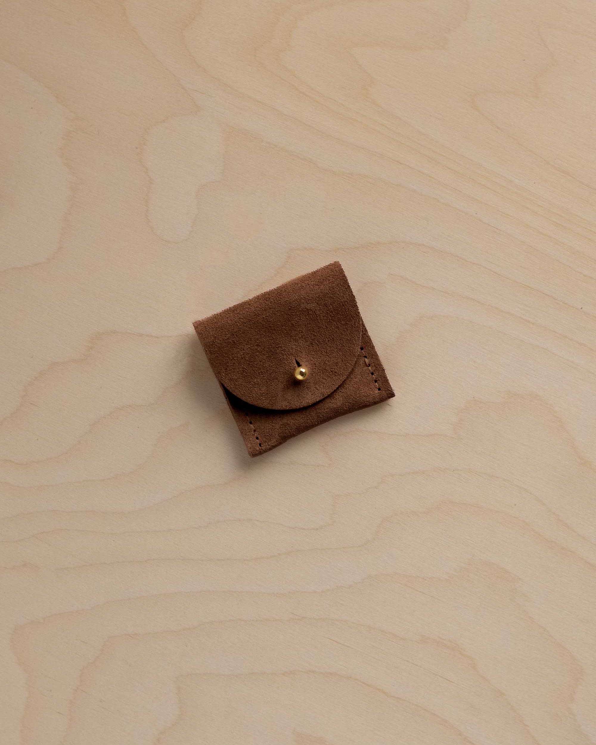 A small brown leather pouch with a flap closure and a gold-tone snap button, placed on a light wood grain surface.