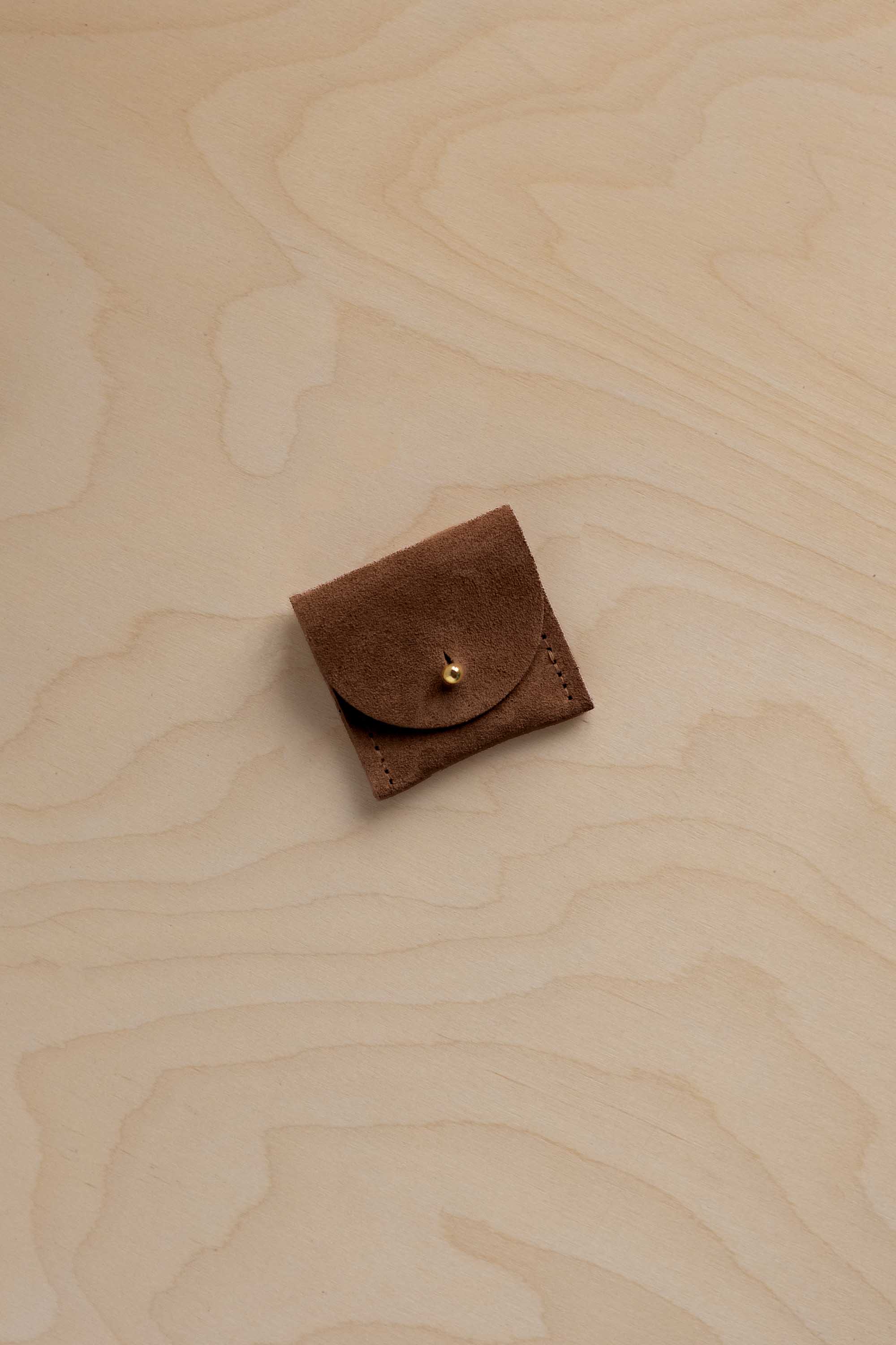 A small brown leather pouch with a flap closure and a gold-tone snap button, placed on a light wood grain surface.