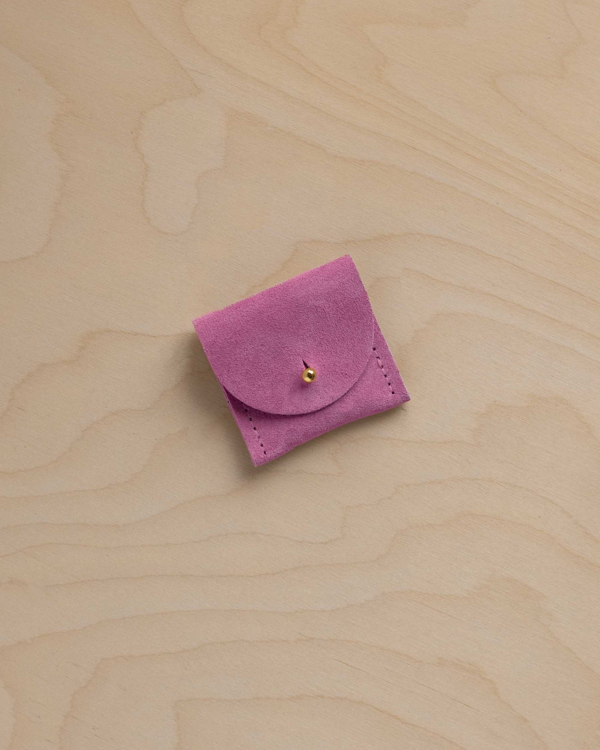 A small light magenta pink suede leather ring pouch with a flap closure and a tiny gold button, placed on a light wood grain surface.