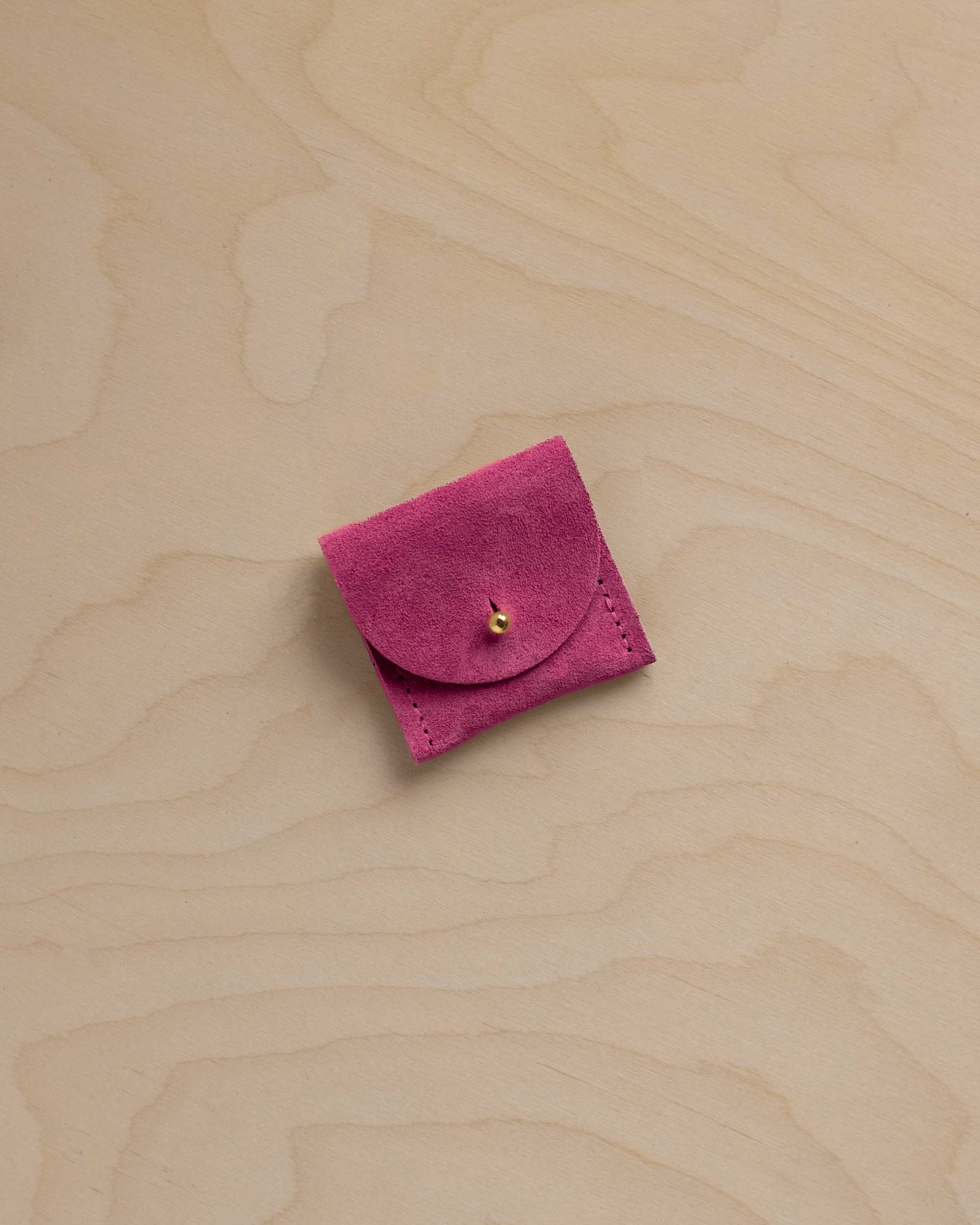 A small magenta pink suede jewelry pouch with a flap closure and a tiny gold bead accent, placed on a light wood grain background.