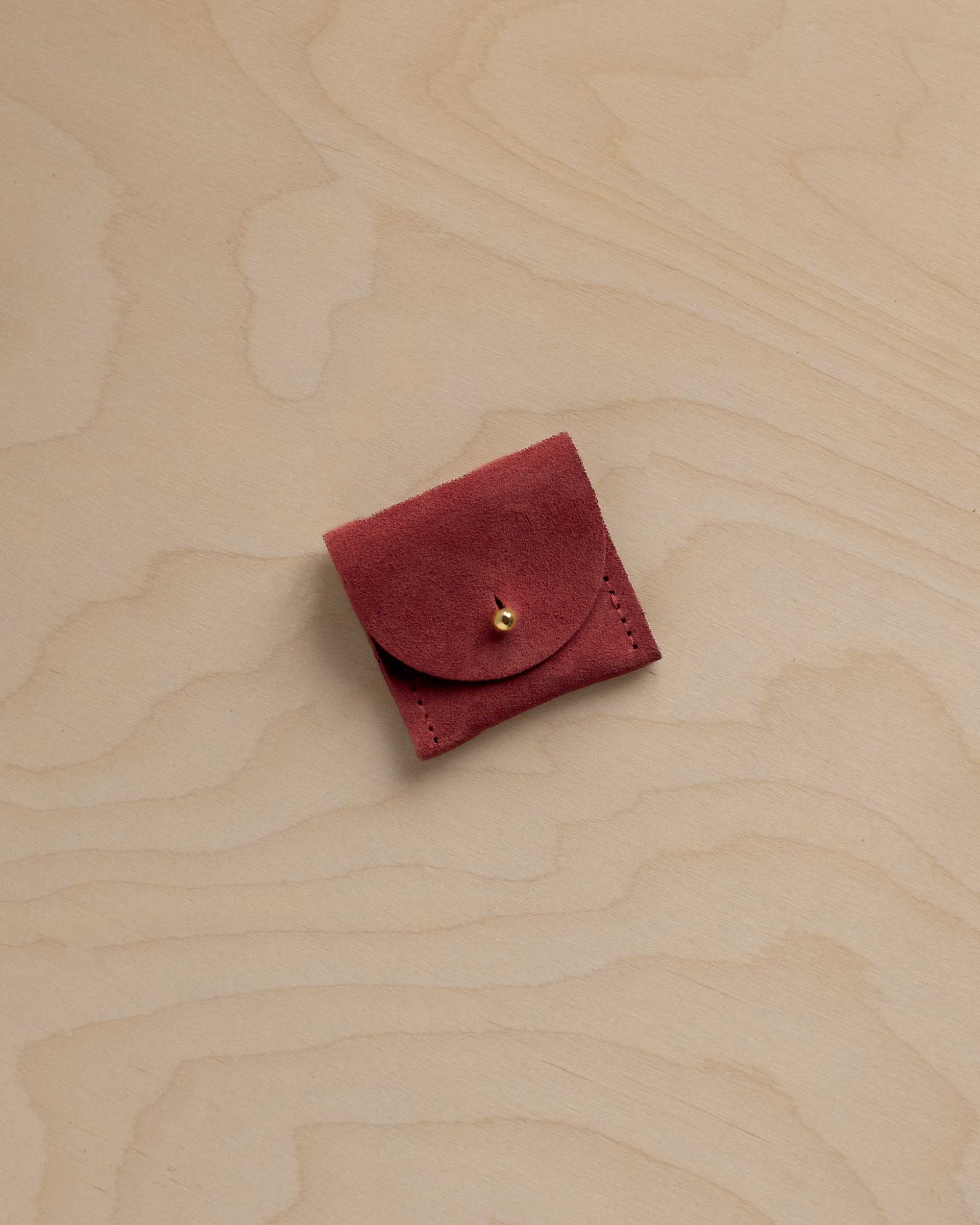 A small rust red suede pouch with a gold button closure, placed on a light wood grain background.