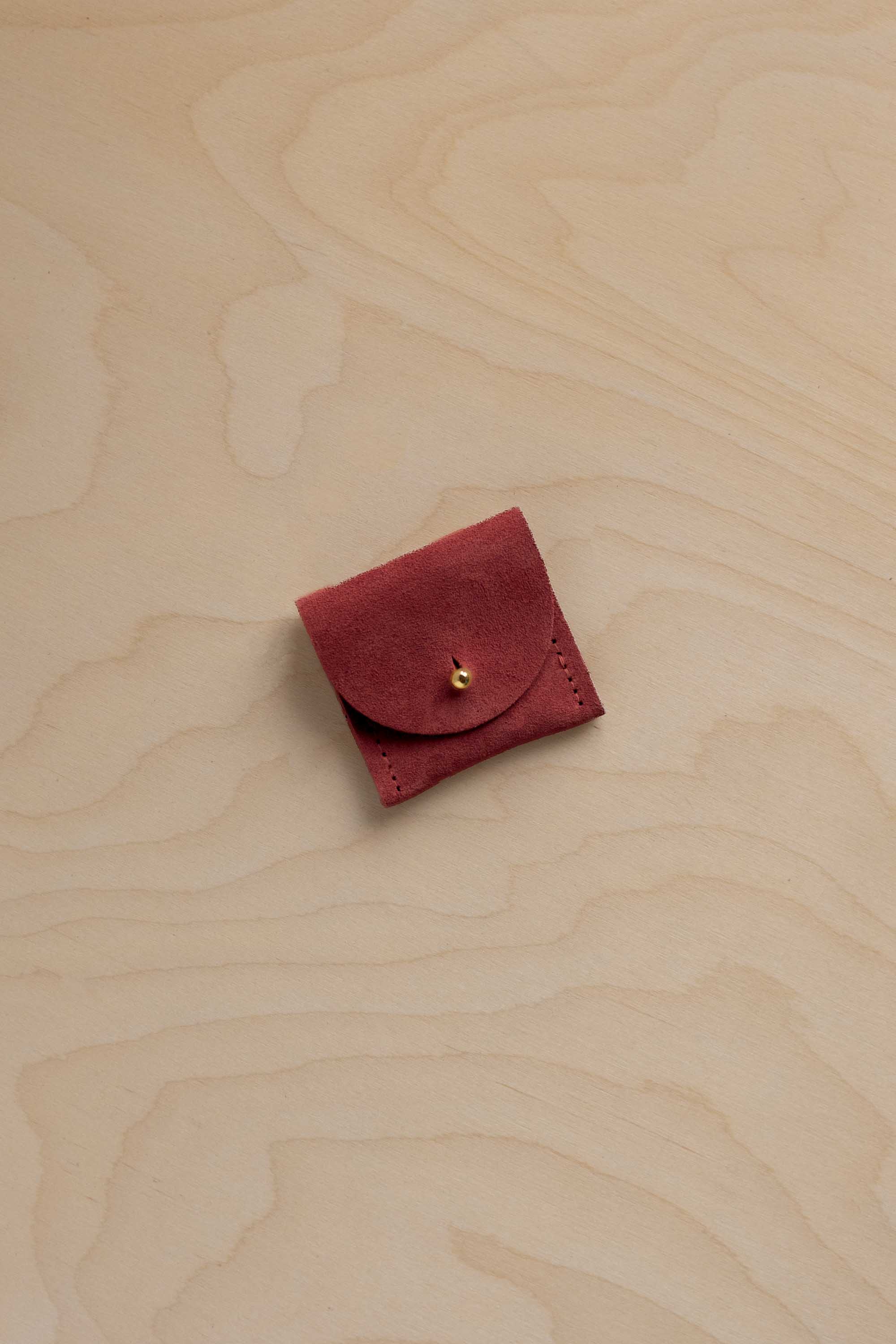 A small rust red suede pouch with a gold button closure, placed on a light wood grain background.