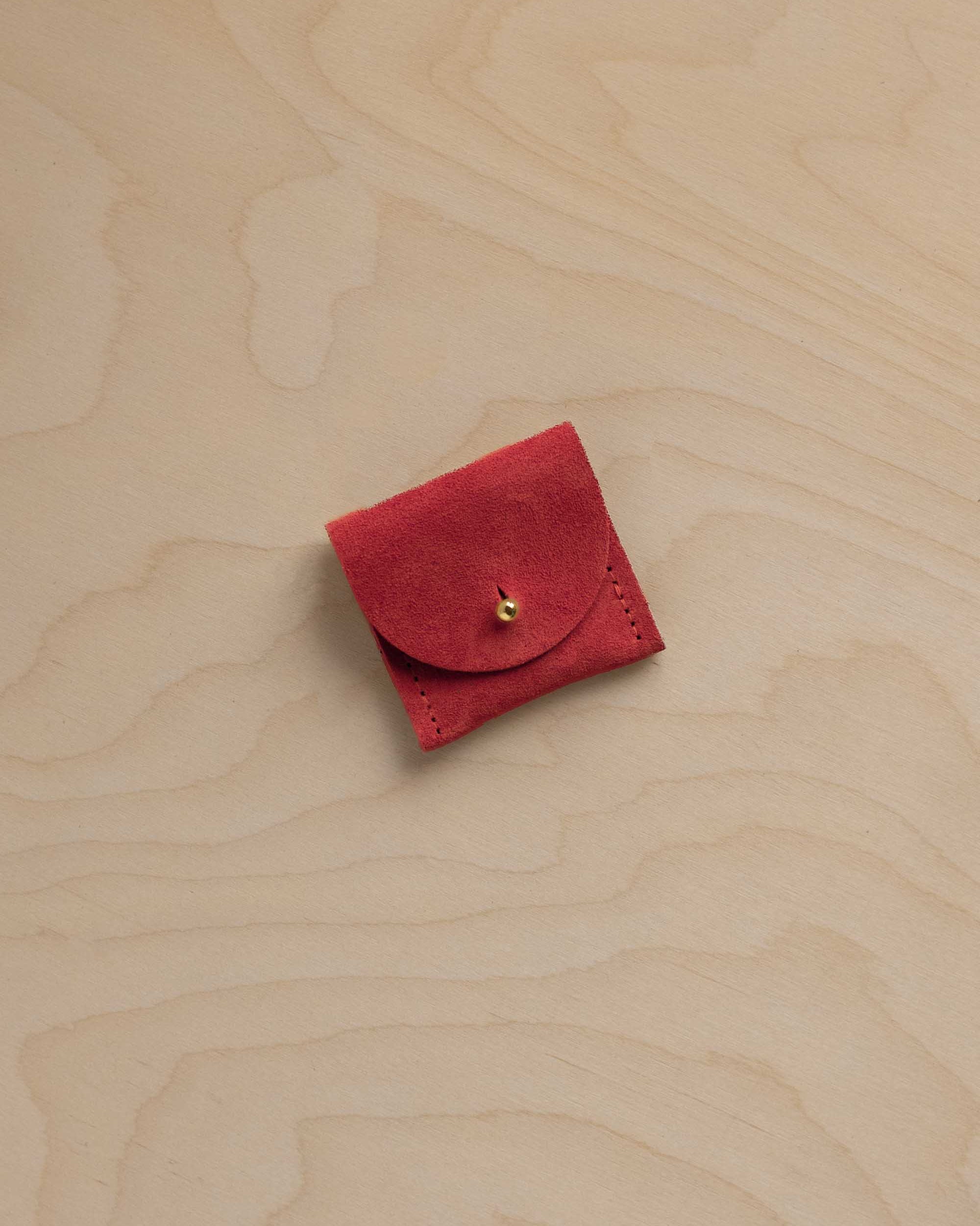 A small red suede jewellery pouch with a circular flap and a tiny gold button, placed on a light wood grain surface.