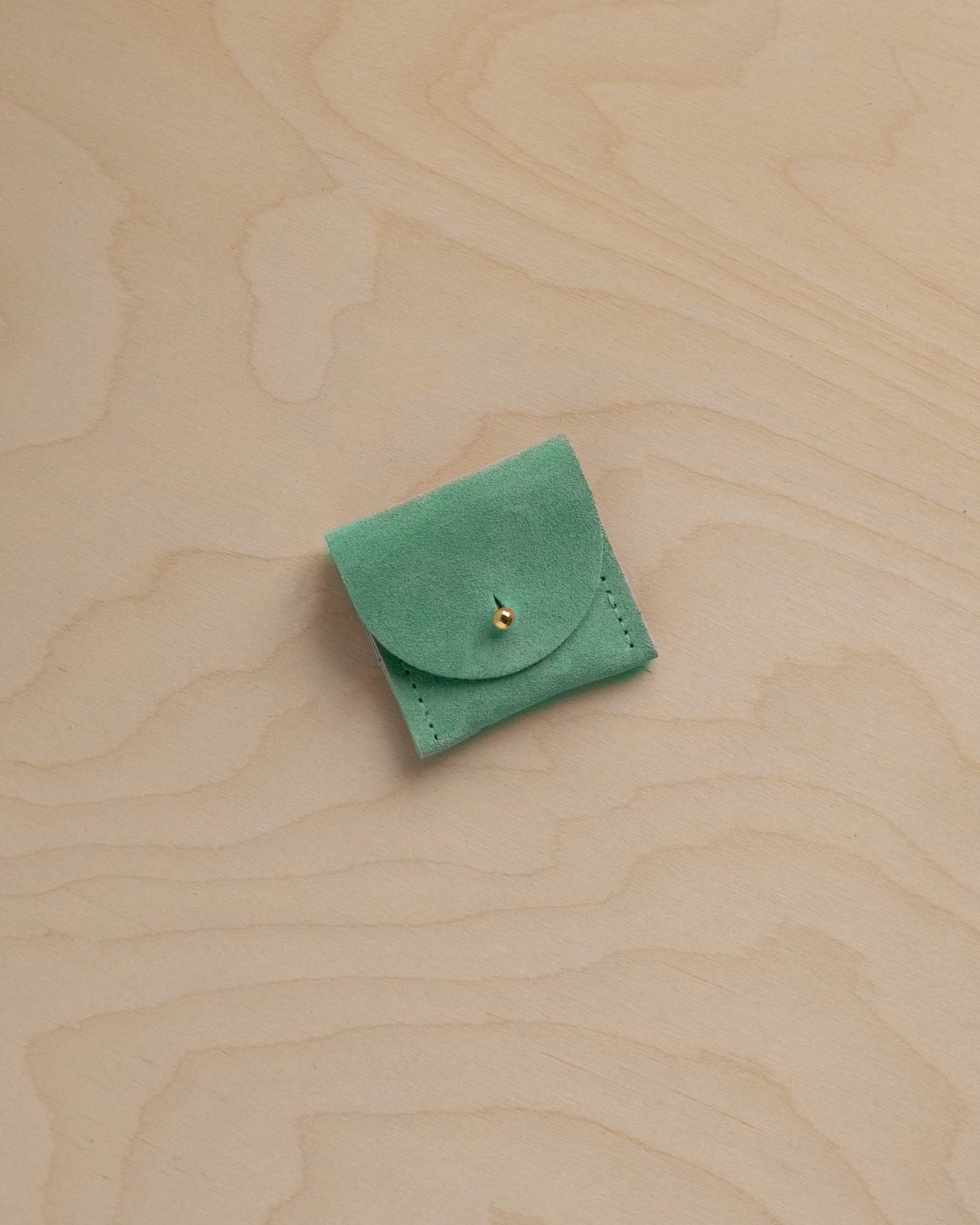 A small light emerald green suede pouch with a flap closure and a tiny gold button, placed on a light beige wood-grain surface.