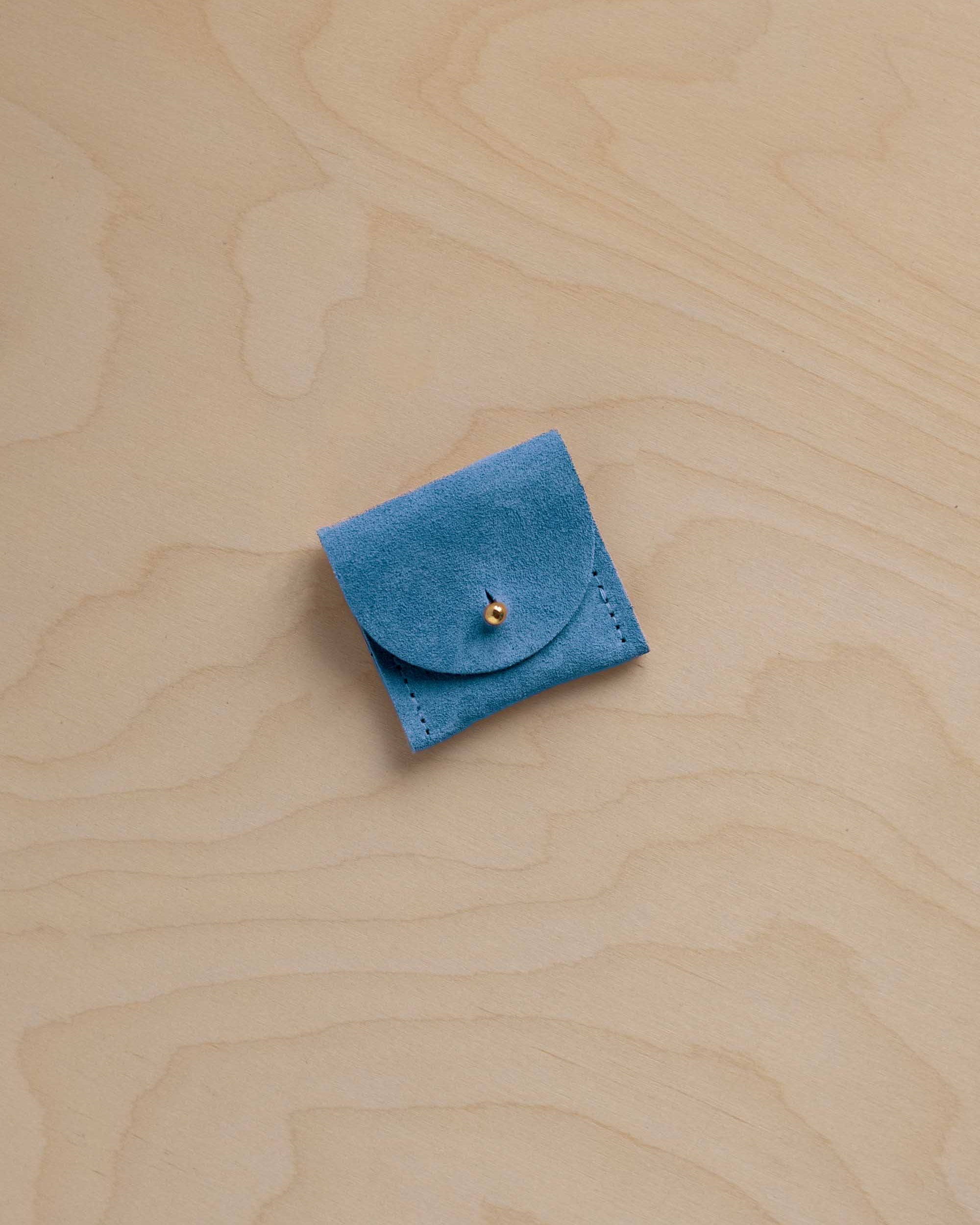 A small bright blue suede leather jewellery pouch with a flap closure and a tiny gold bead accent, placed on a light wood grain surface.