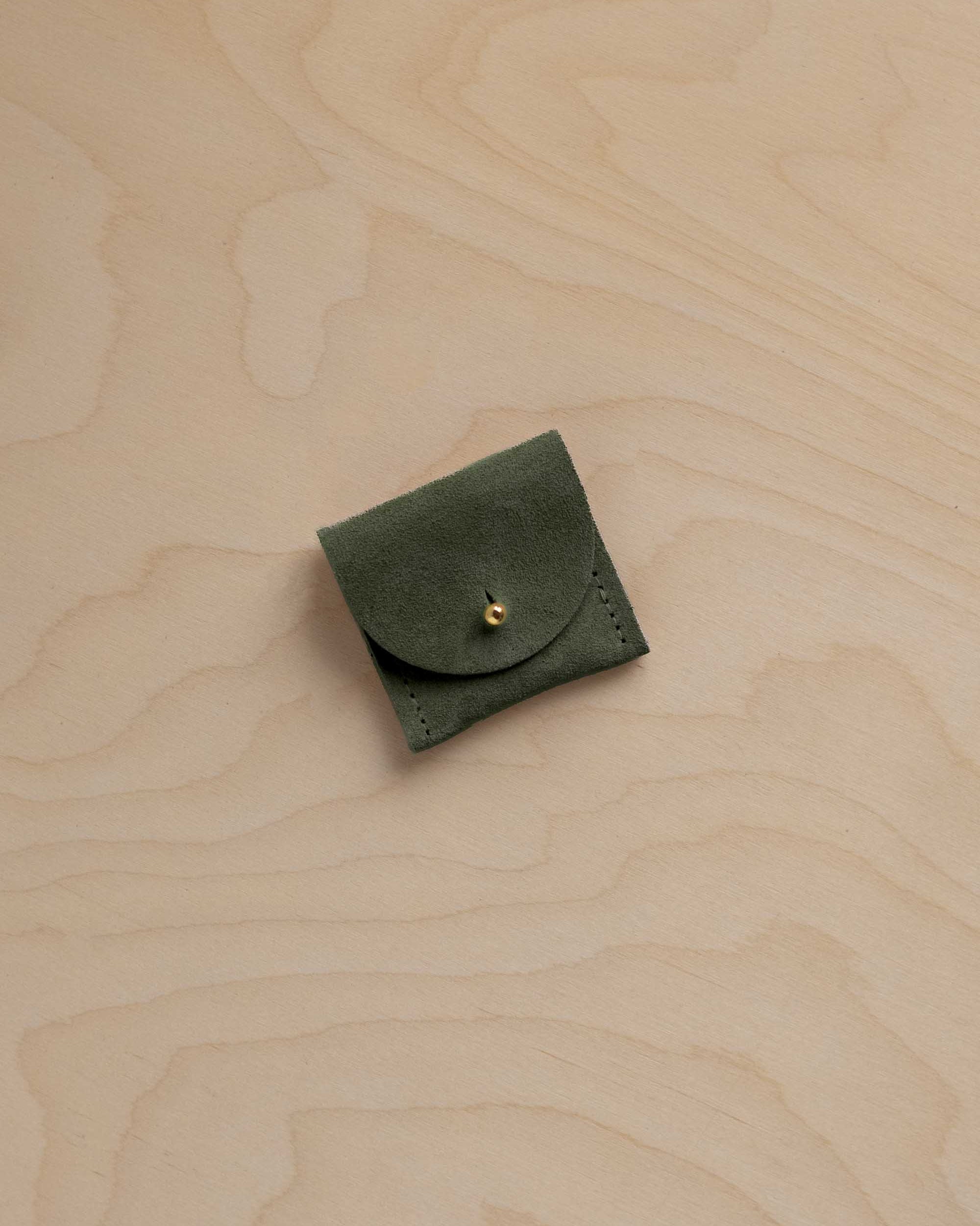 A small olive green leather pouch with a flap closure and a gold-tone snap button, placed on a light wood grain surface.