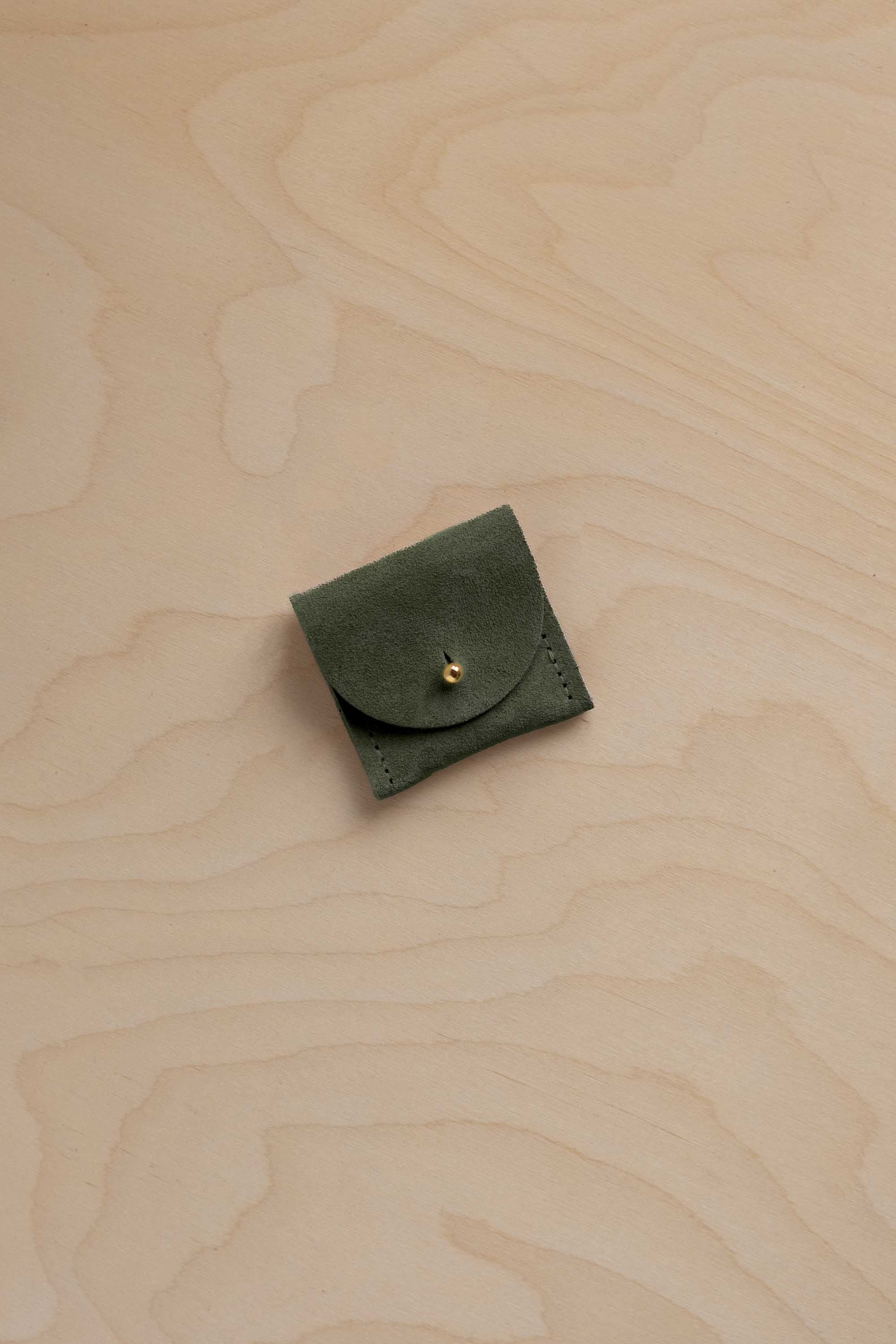 A small olive green leather pouch with a flap closure and a gold-tone snap button, placed on a light wood grain surface.
