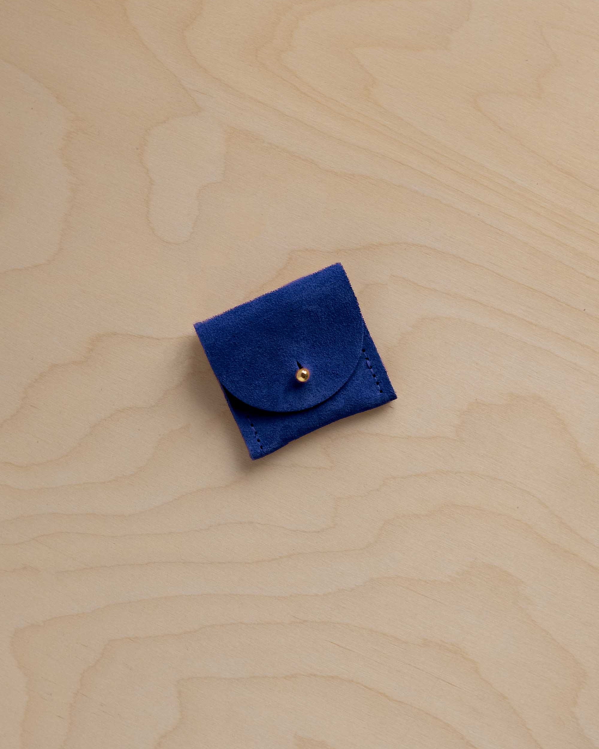 A small square dark blue suede pouch with a flap closure and a single gold button, placed on a light wood grain surface.