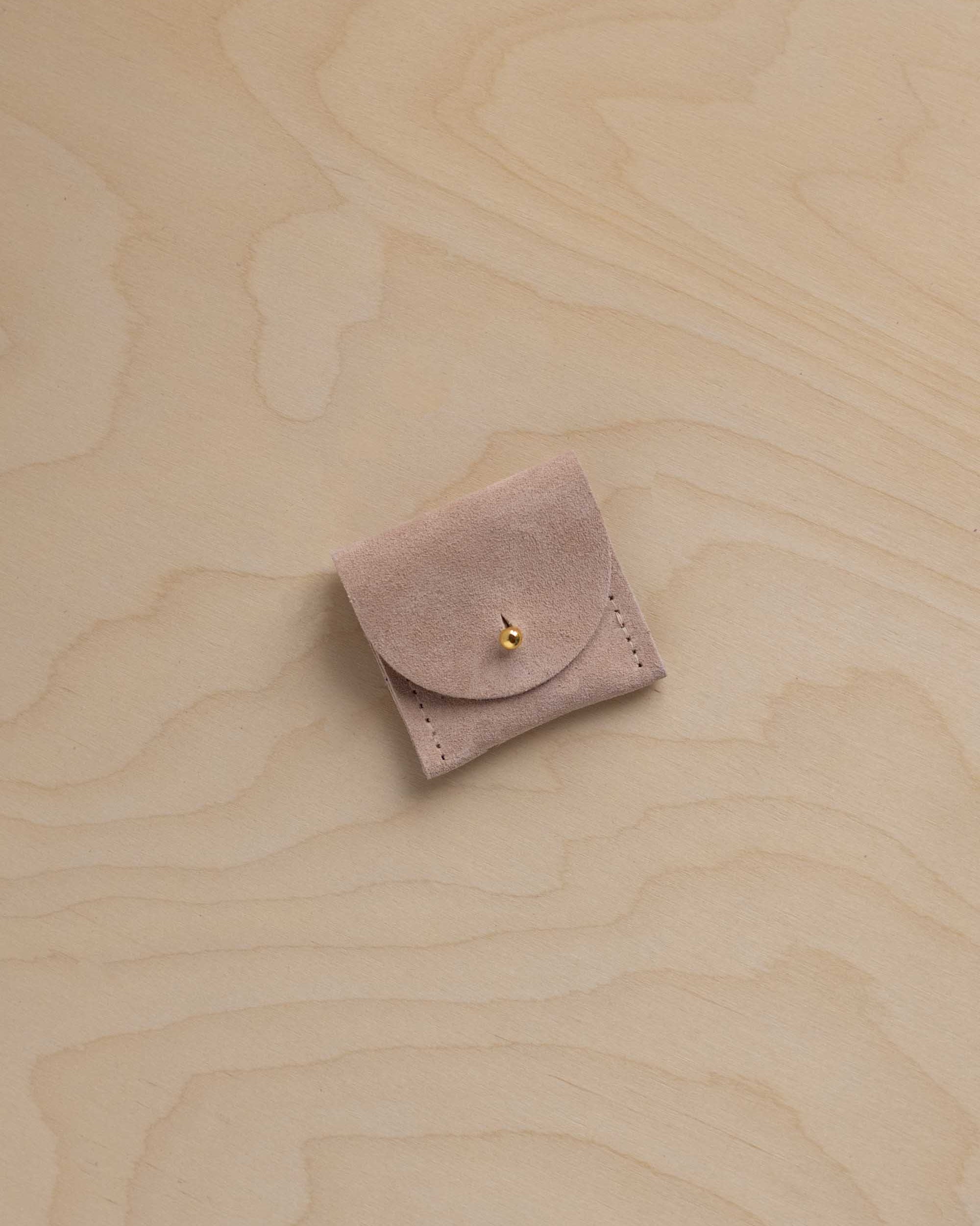 A small dusty beige / pink suede jewelry pouch with a gold button, placed on a light wood grain background.