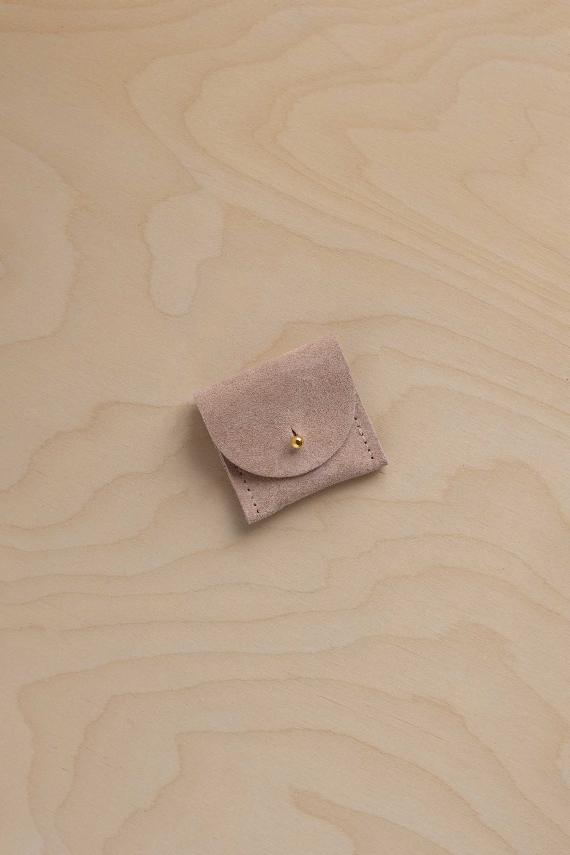 A small dusty beige / pink suede jewelry pouch with a gold button, placed on a light wood grain background.
